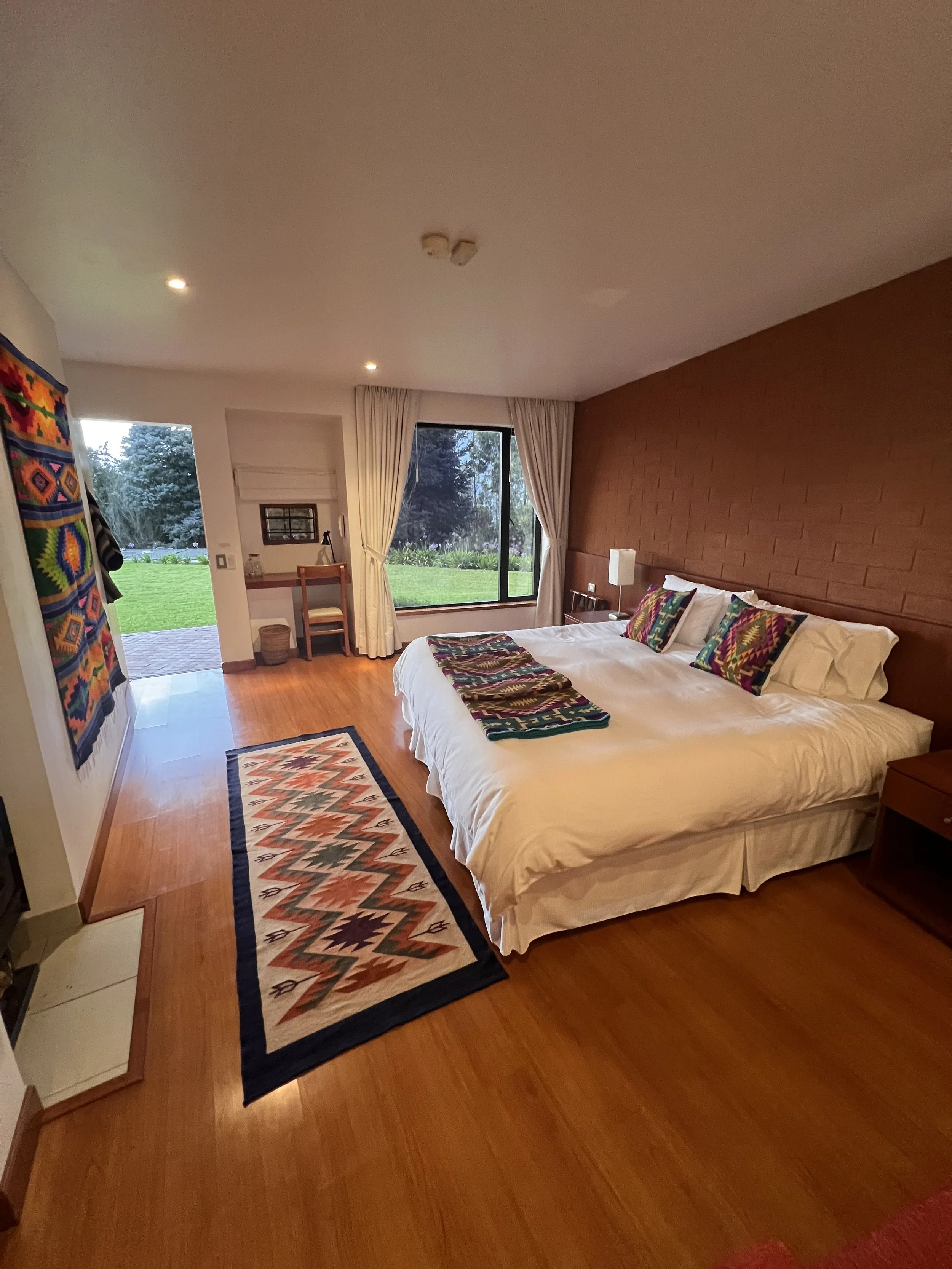 A cozy bedroom with a large bed adorned with colorful, patterned pillows and a matching throw. The room has a wooden floor, a patterned runner rug at the foot of the bed, and a window with cream curtains revealing a green outdoor view. There is a small nightstand with a lamp and other minimal decor, a doorway leading outside, and a vibrant woven wall hanging on the left side of the room.