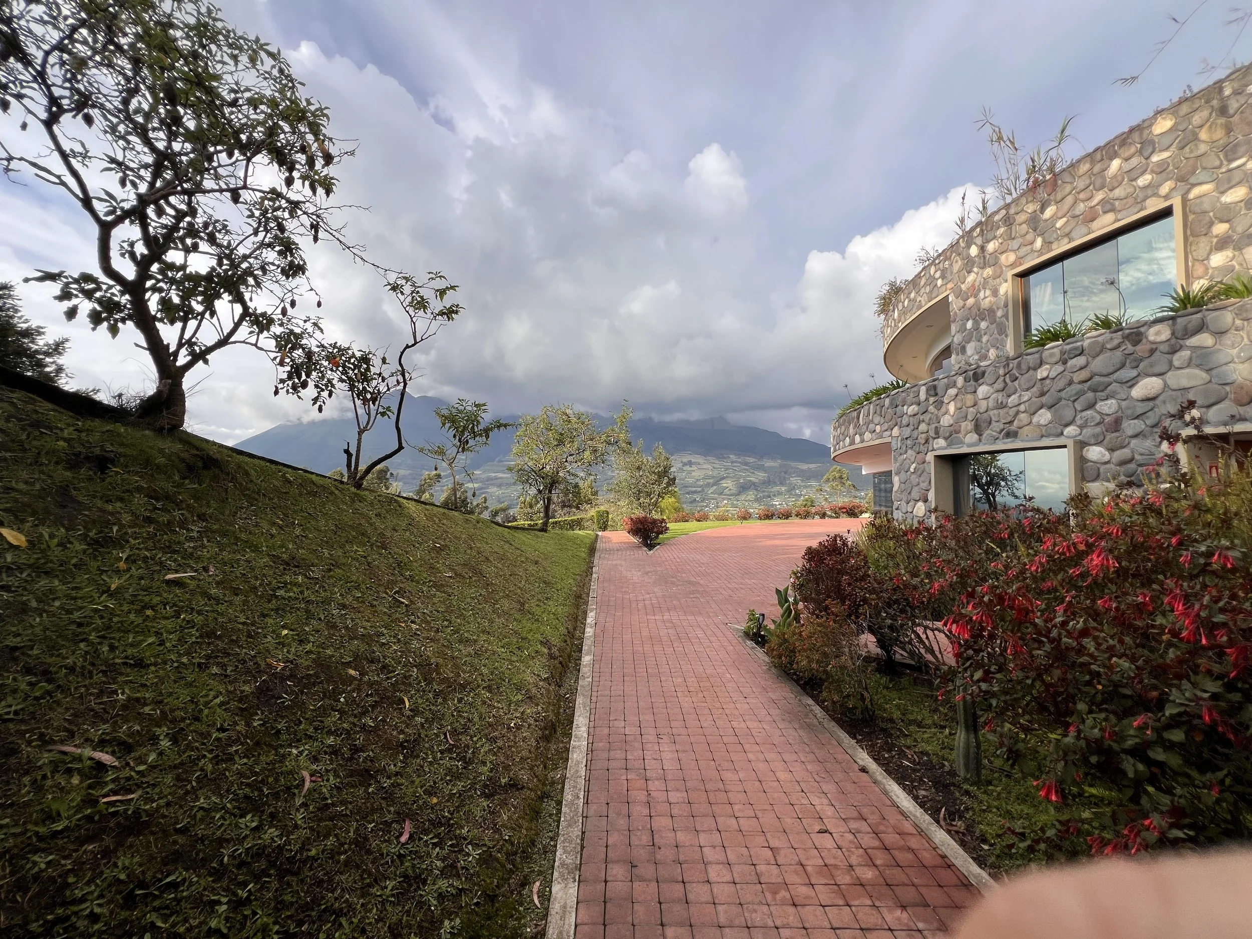 A scenic view of a modern stone building with large windows, a paved walkway, trees, and a mountain in the background under a partly cloudy sky.