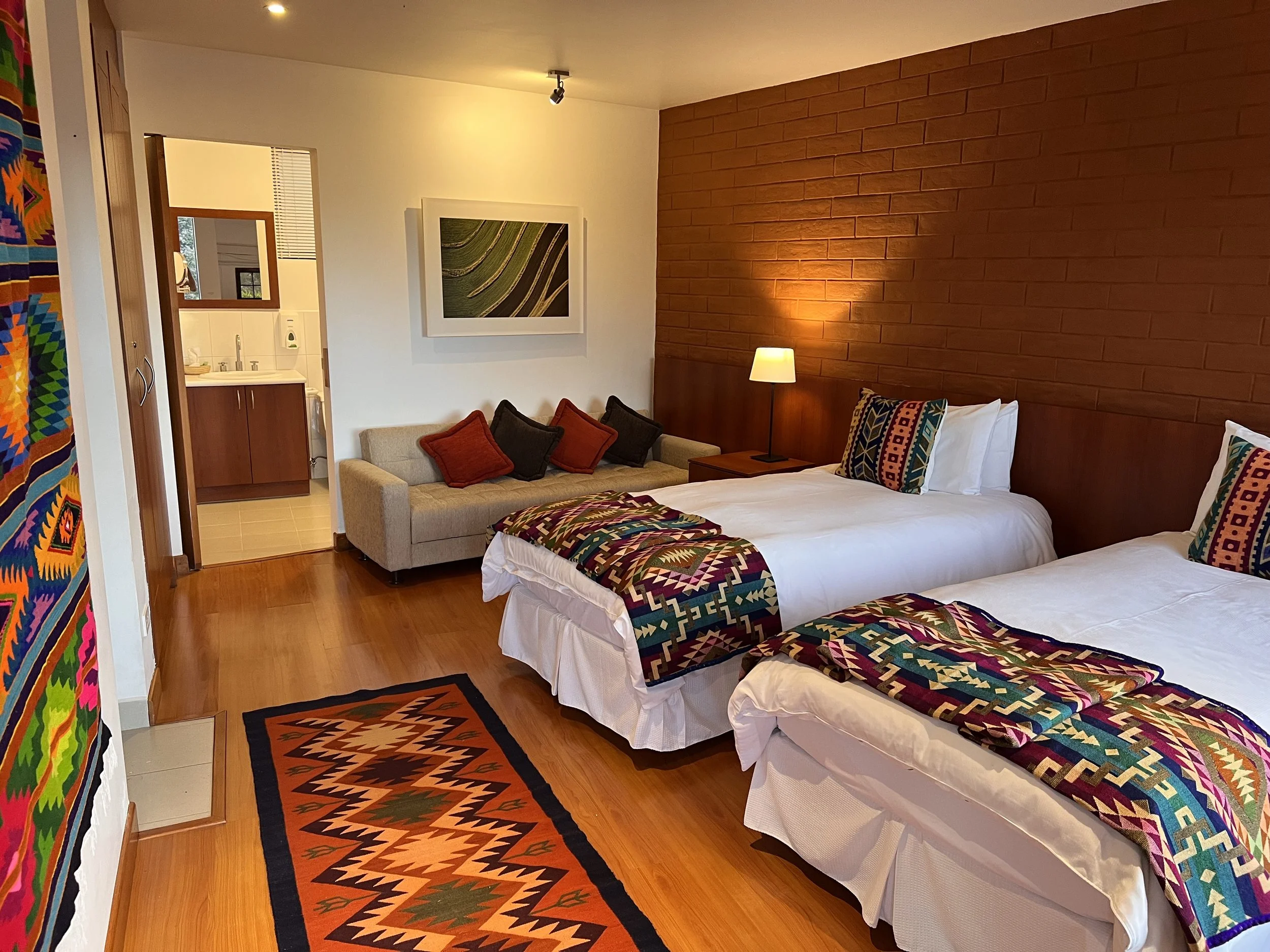 A hotel room with two neatly made beds covered with colorful patterned blankets, a small sofa with red and black cushions, a wooden nightstand with a lamp, a brick accent wall behind the bed, and a painting above the sofa. An orange and black patterned rug is on the wooden floor.