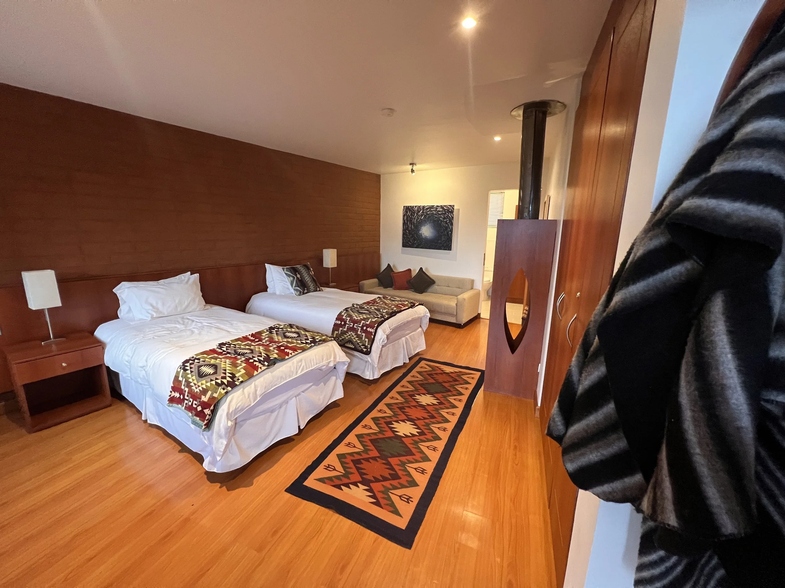 A hotel room with two single beds covered with colorful blankets, a beige sofa with black and red pillows, a wooden headboard, and two white lamps on bedside tables. There is a patterned rug on the wooden floor and a piece of abstract artwork on the wall. A portion of a person's arm and coat is visible in the right foreground.