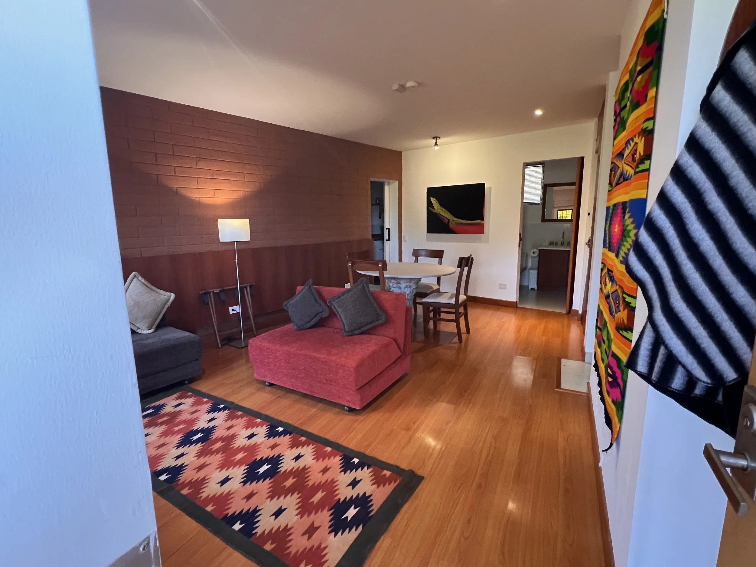 Living room with red brick accent wall, round dining table with chairs, a red sofa with gray cushions, a colorful patterned rug, a white floor lamp, and artwork on the walls.