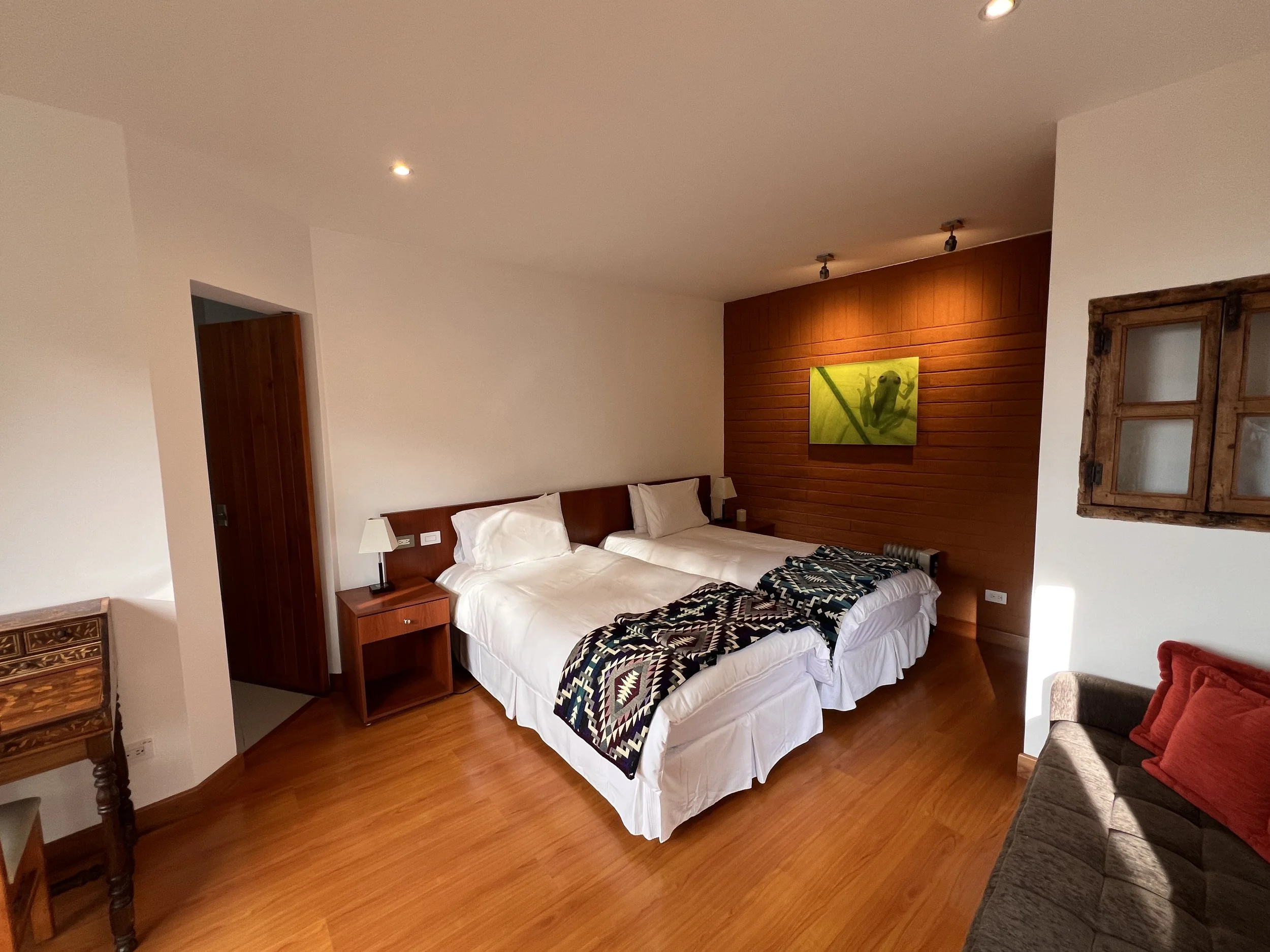 A bedroom with two single beds with white bedding, wooden headboards, and colorful geometric-patterned quilts. There are two bedside tables with lamps, and a wooden wall with a green frog painting. An open wooden window frame is on the wall to the right.