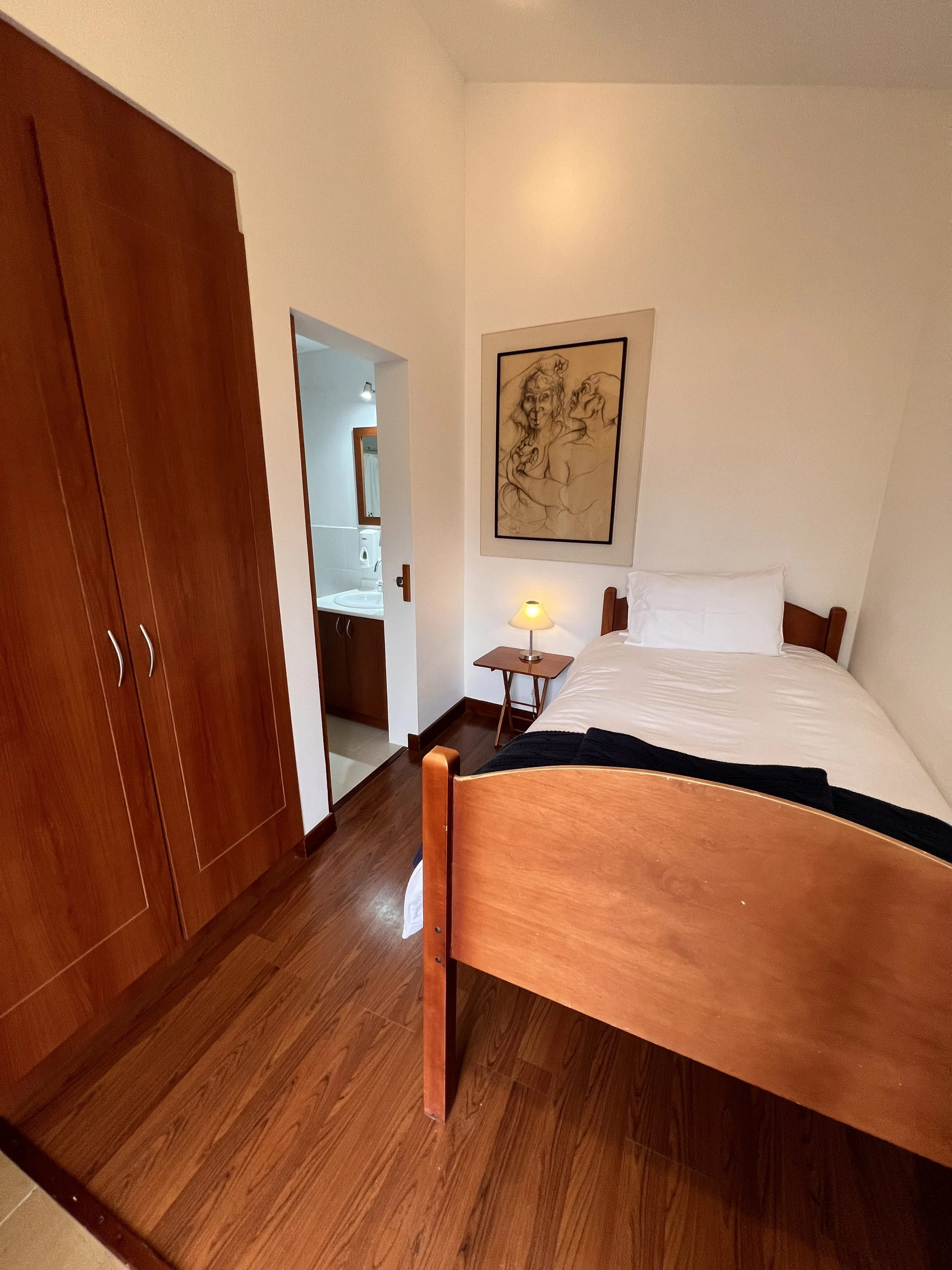 A hotel room with a bed, nightstand, lamp, framed artwork, and a closet with wooden doors. There's a bathroom visible in the background.