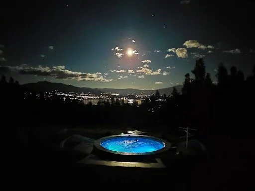 Nighttime view of a hot tub outdoors under a moonlit sky with clouds, trees, and distant city lights.