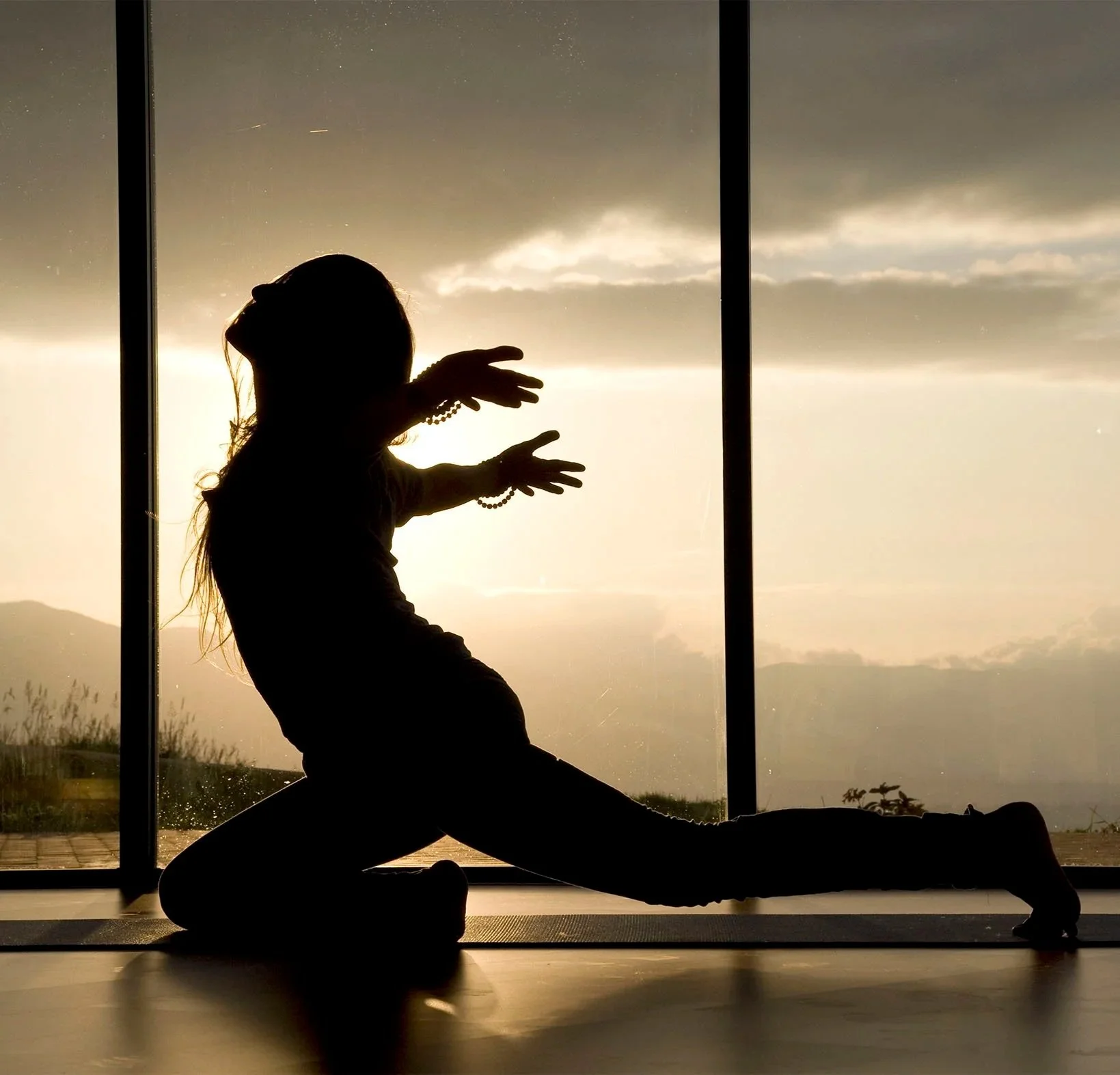 Silhouette of a person performing a stretching exercise on a yoga mat, with large windows showing a sunset or sunrise and mountains and clouds in the background.