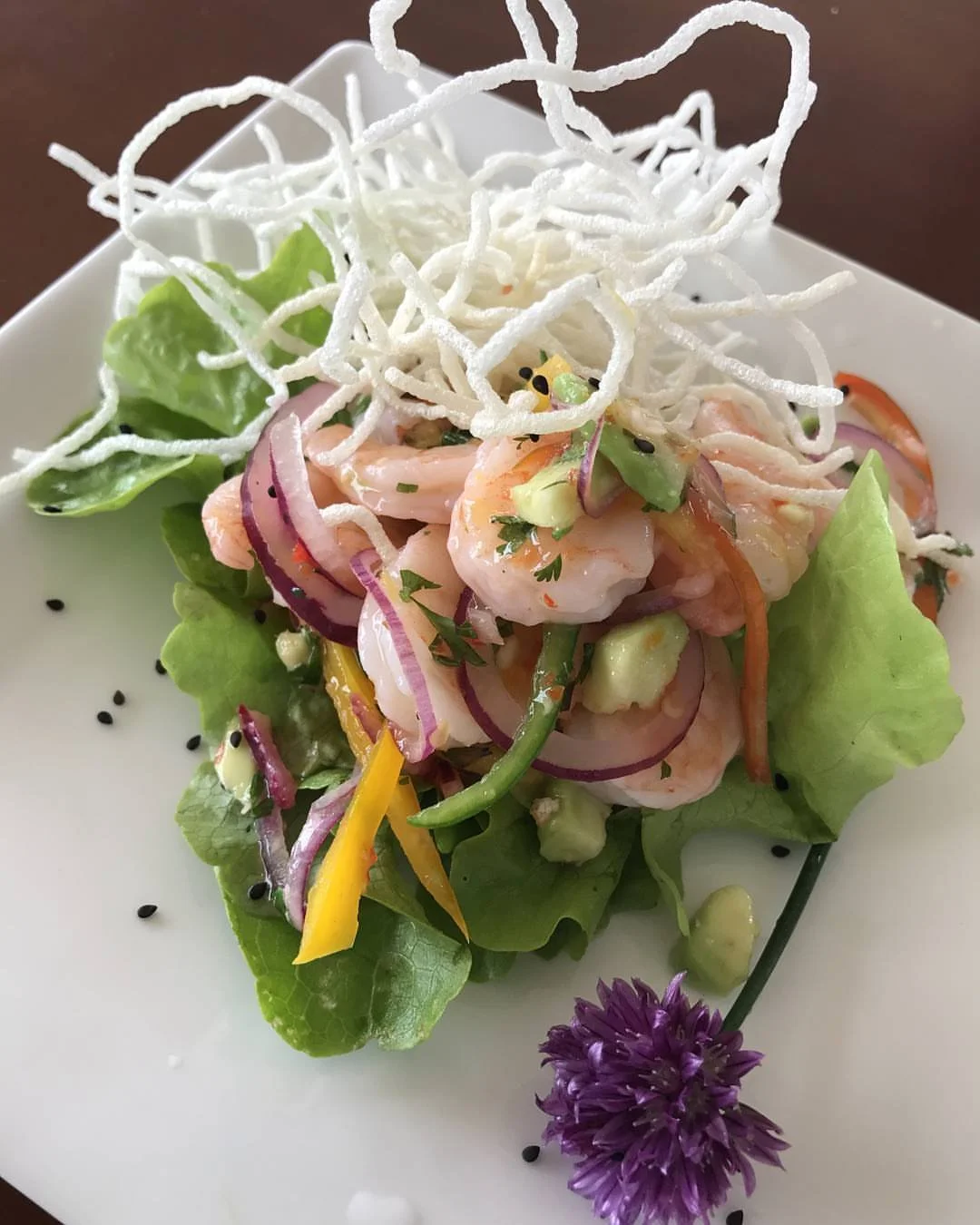 Fresh seafood salad with shrimp, sliced red onions, yellow bell peppers, lettuce, cucumber, and crispy rice noodles, garnished with black sesame seeds and a purple edible flower.