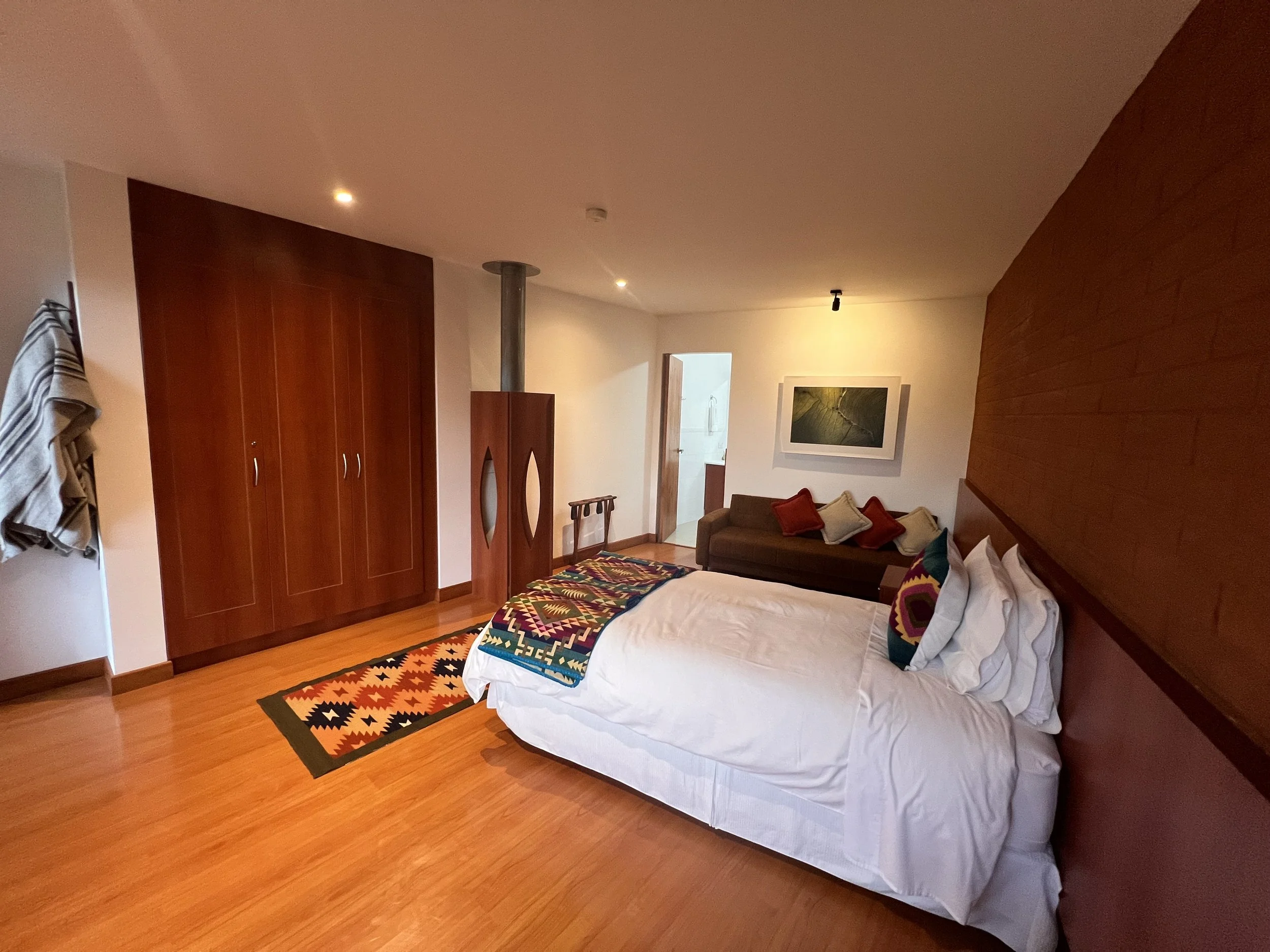 A bedroom with hardwood floors, a bed with white bedding and colorful pillows, a wooden wardrobe, a colorful geometric rug, a sofa with pillows, a framed picture on the wall, and partially visible clothes hanging on a rack.