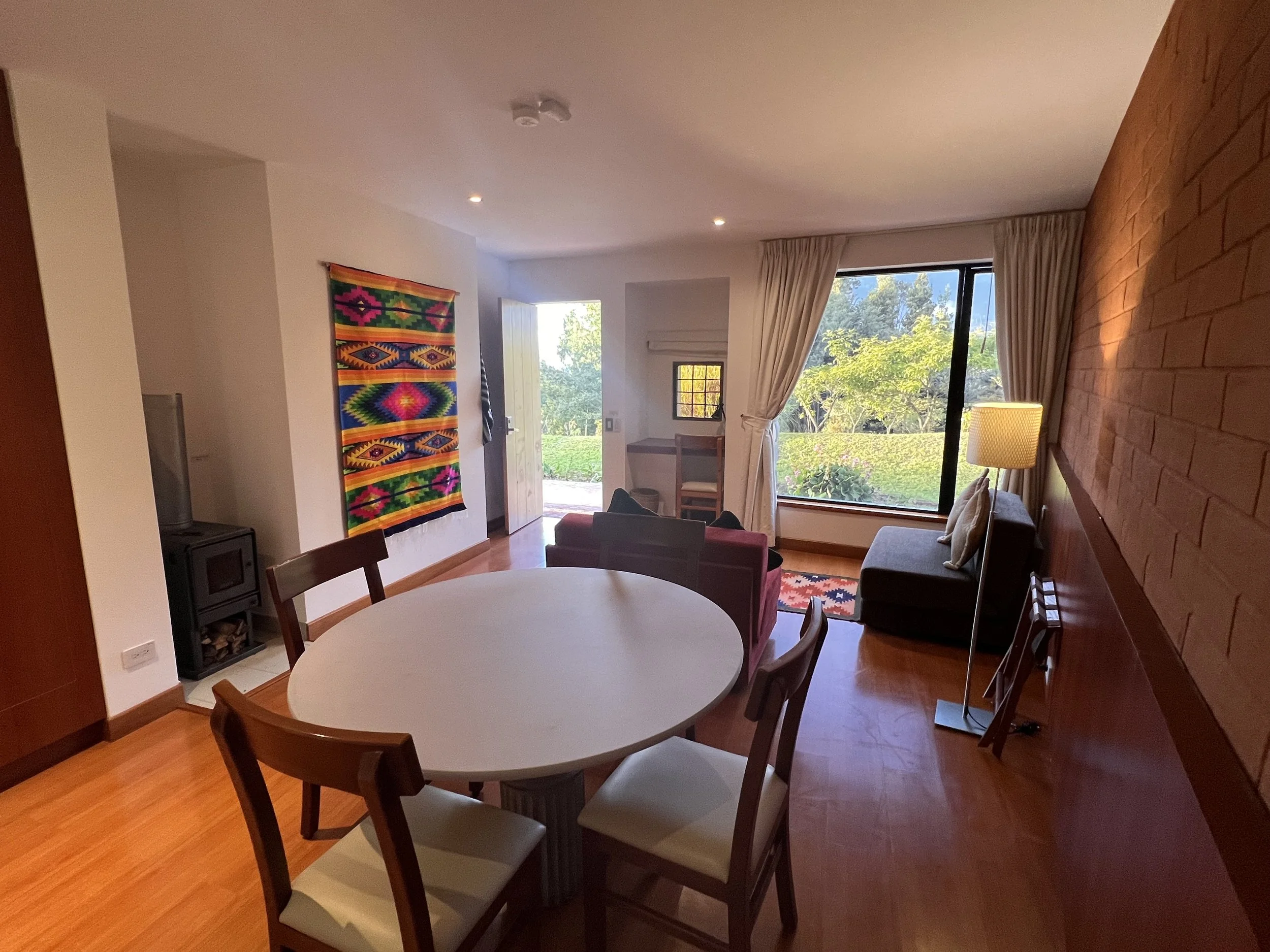 The interior of a living room with a round dining table and chairs, a window with curtains, a brick accent wall, a colorful wall hanging, and a door leading outside with visible greenery.