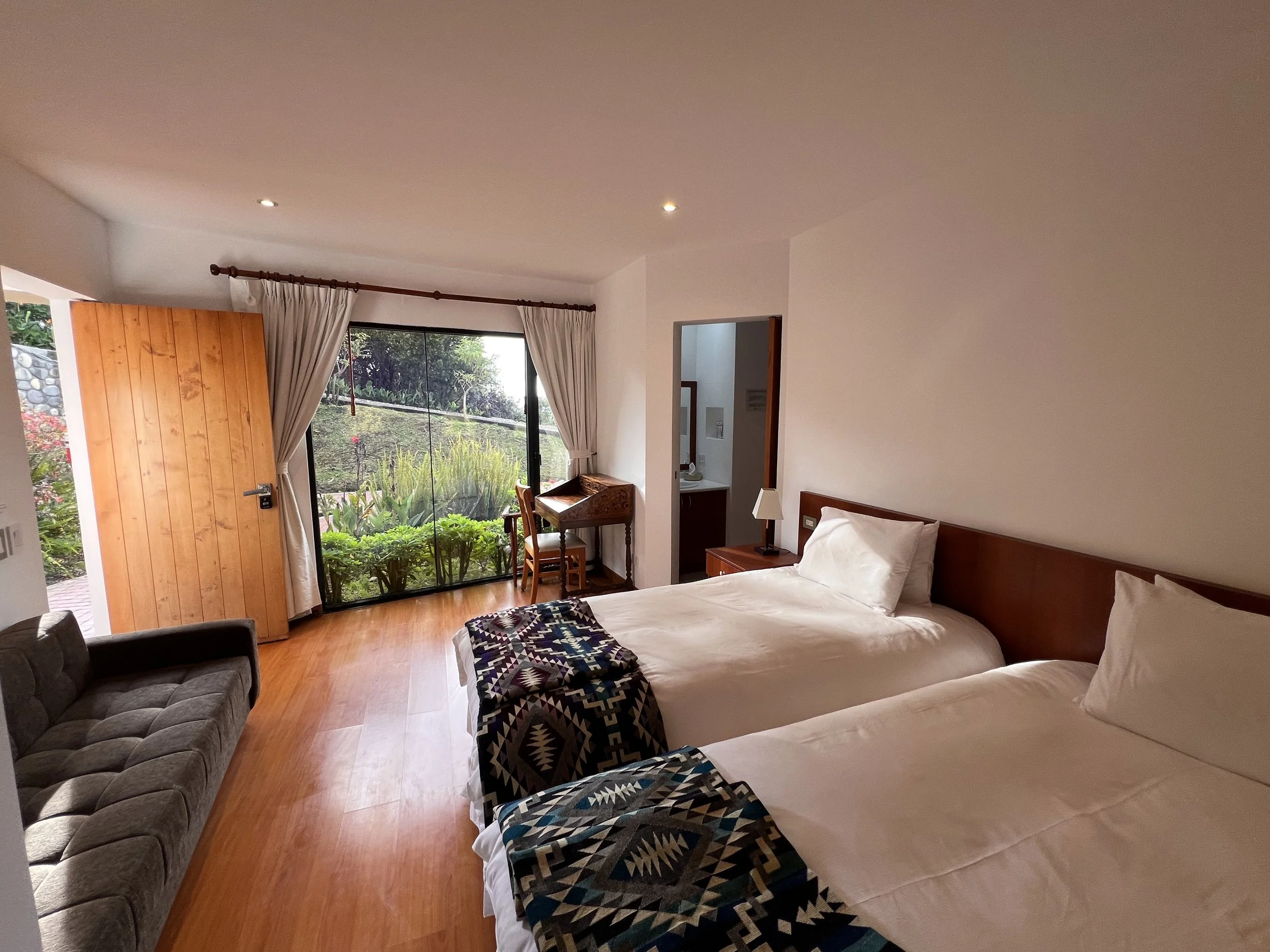 A bedroom with two beds, a wooden door, large windows with curtains, and a sliding glass door leading to a garden outside.