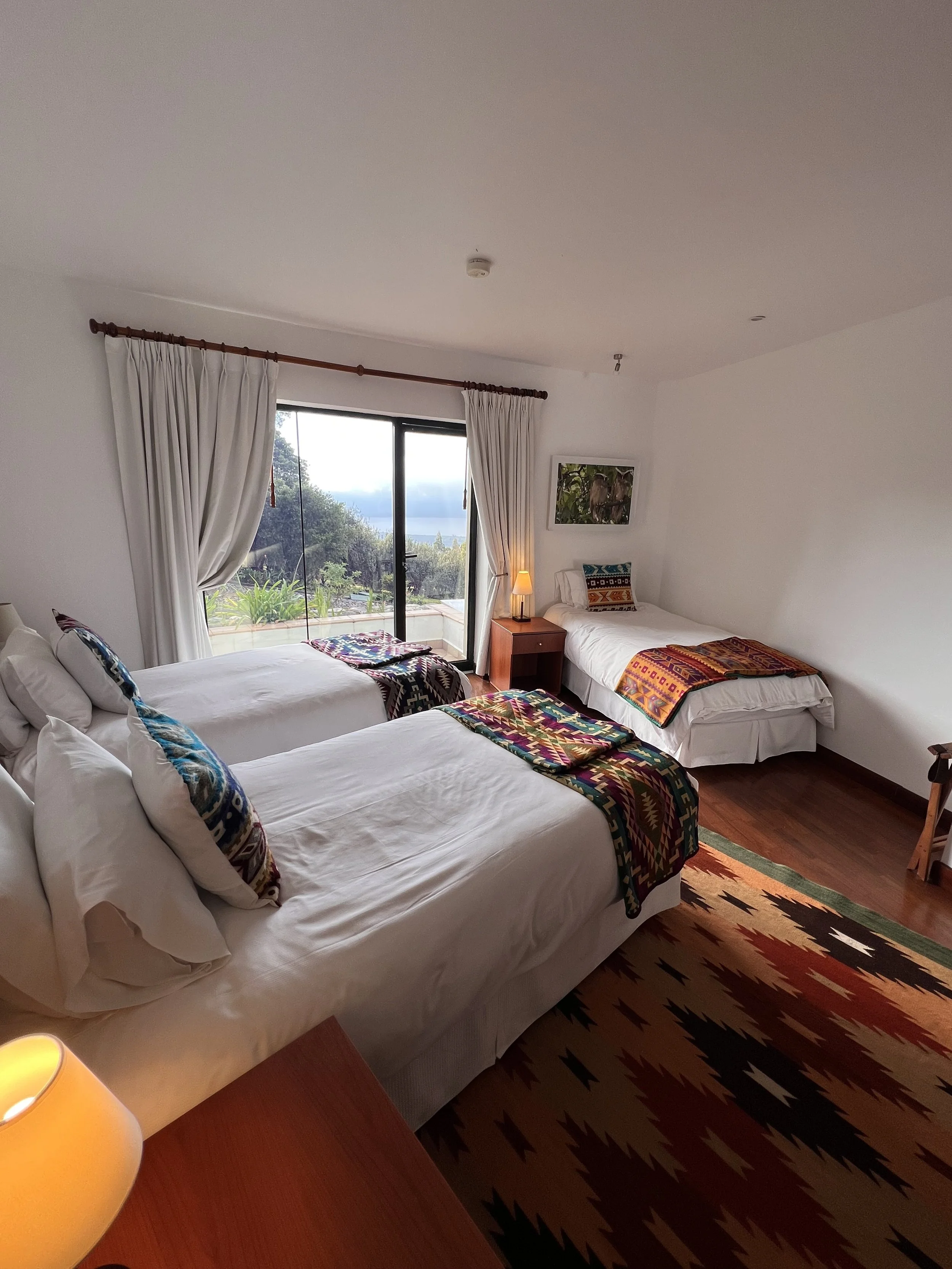 A bedroom with three beds, decorated with colorful patterned blankets and pillows. A large window with white curtains offers a view of greenery and trees outside. There is a wooden nightstand with a small lamp between two beds, and a framed picture on the wall above.