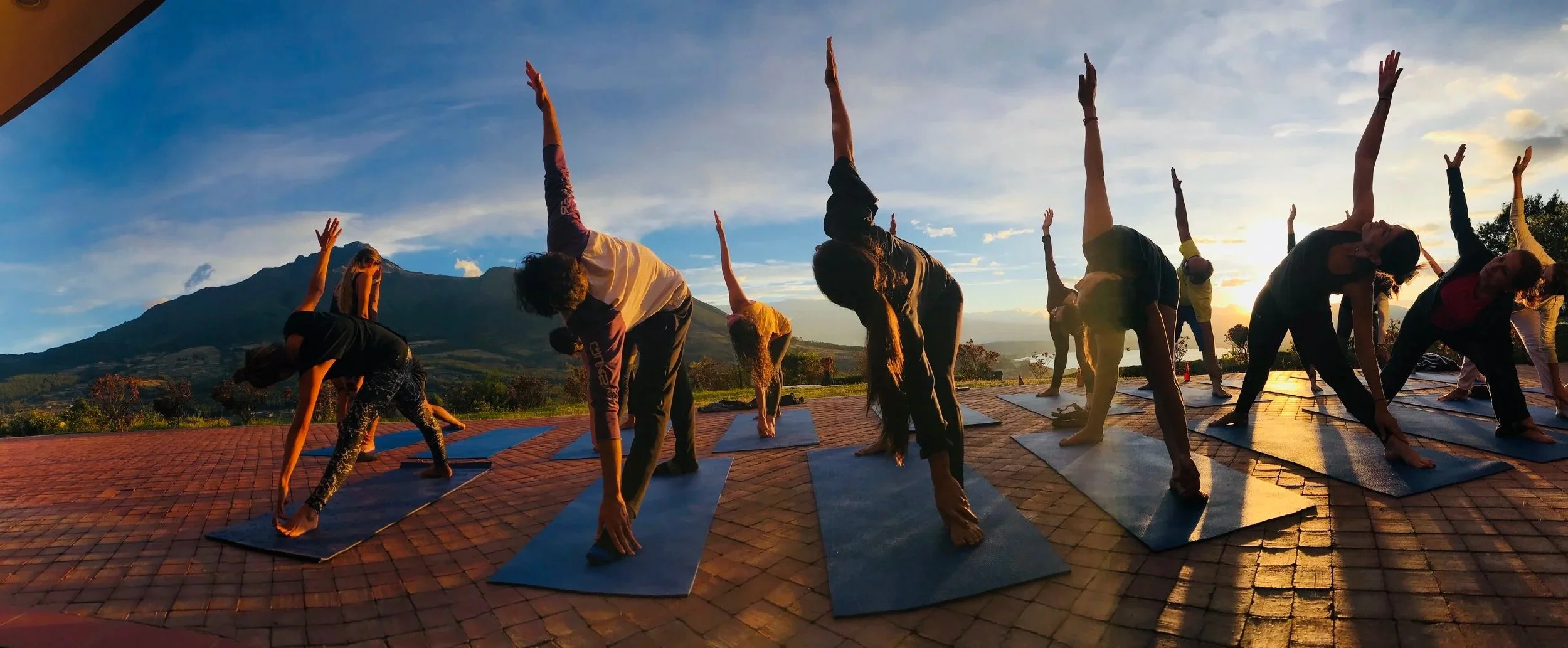 People practicing yoga outdoors on mats during sunrise or sunset with mountains and a scenic landscape in the background.
