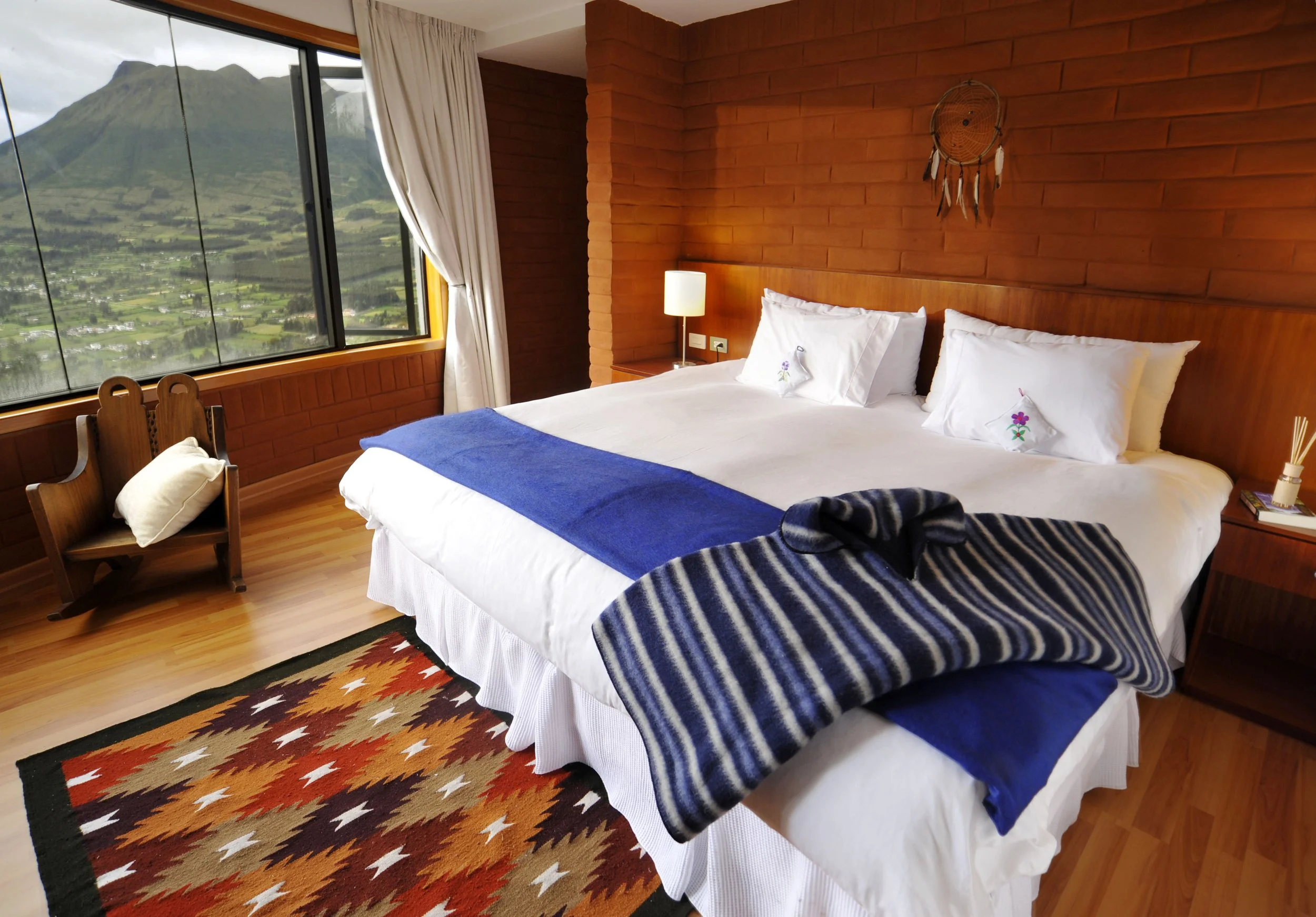 A cozy bedroom with a large window showcasing a scenic mountain view. The room features a bed with white linens, a blue runner at the foot, and decorative pillows. There's a wooden side table with a lamp, a wooden chair with a pillow, a colorful patterned rug, and a dreamcatcher hanging on a brick wall.