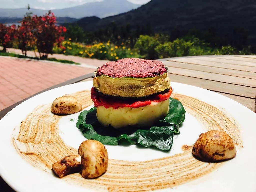 Elegant dish with layered vegetables, including eggplant, tomato, and zucchini, garnished with spinach and surrounded by mushrooms, served on a white plate outdoors with mountains and greenery in the background.