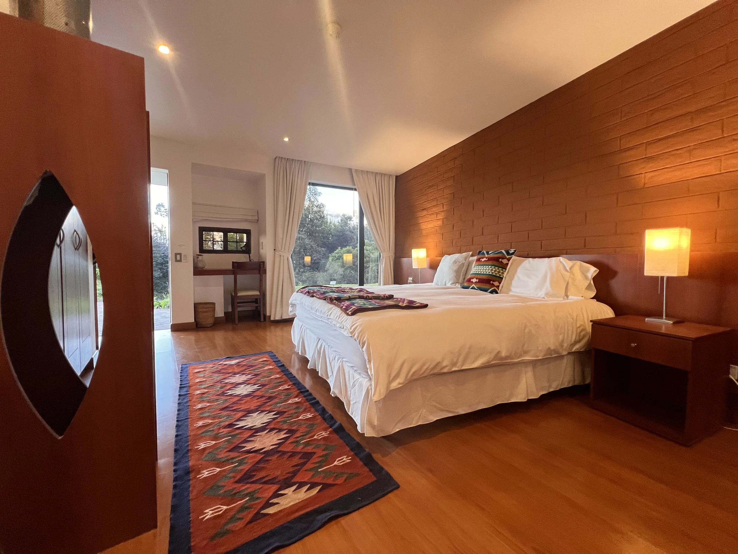 A bedroom with a bed, colorful pillows, a wooden nightstand with a lamp, large windows with curtains, and a patterned rug on wooden floor.