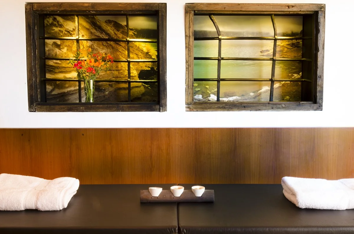 Two landscape posters in rustic wooden frames on a white wall, depicting natural scenery with water and cliffs. A vase with red and pink flowers is in front of the left poster. Below, there is a dark surface with two rolled white towels and three small bowls on a wooden tray.