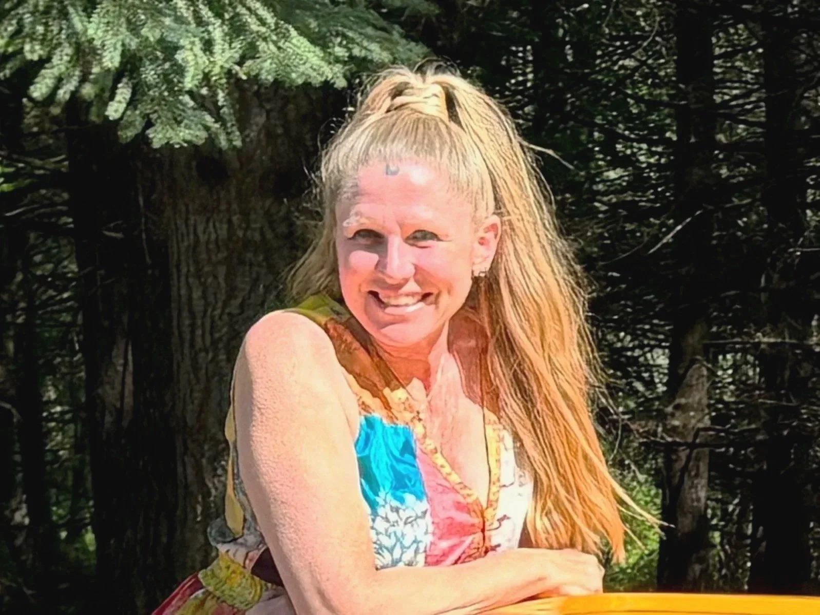A smiling Ayurvedic Consultant with long, light blonde hair in a high ponytail, wearing a colorful sleeveless top, outdoors with trees and greenery in the background.