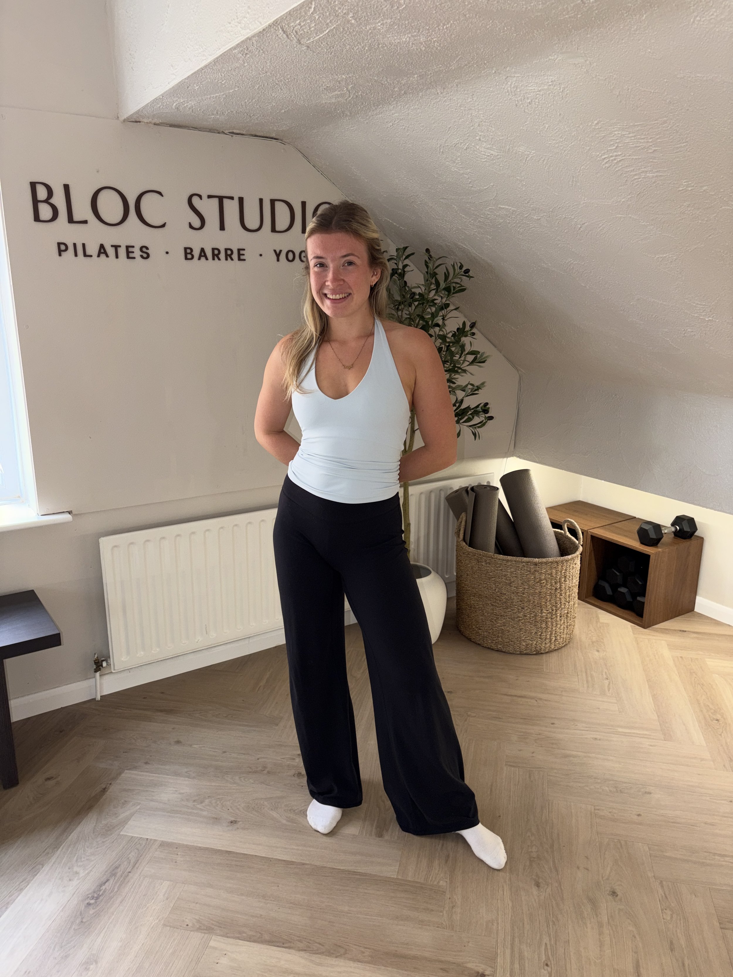 A young woman with long blonde hair smiling at the camera while sitting on a Pilates reformer machine indoors near large windows with natural light. The room has white walls, decorative vases with branches, and a view of a brick building and greenery outside.
