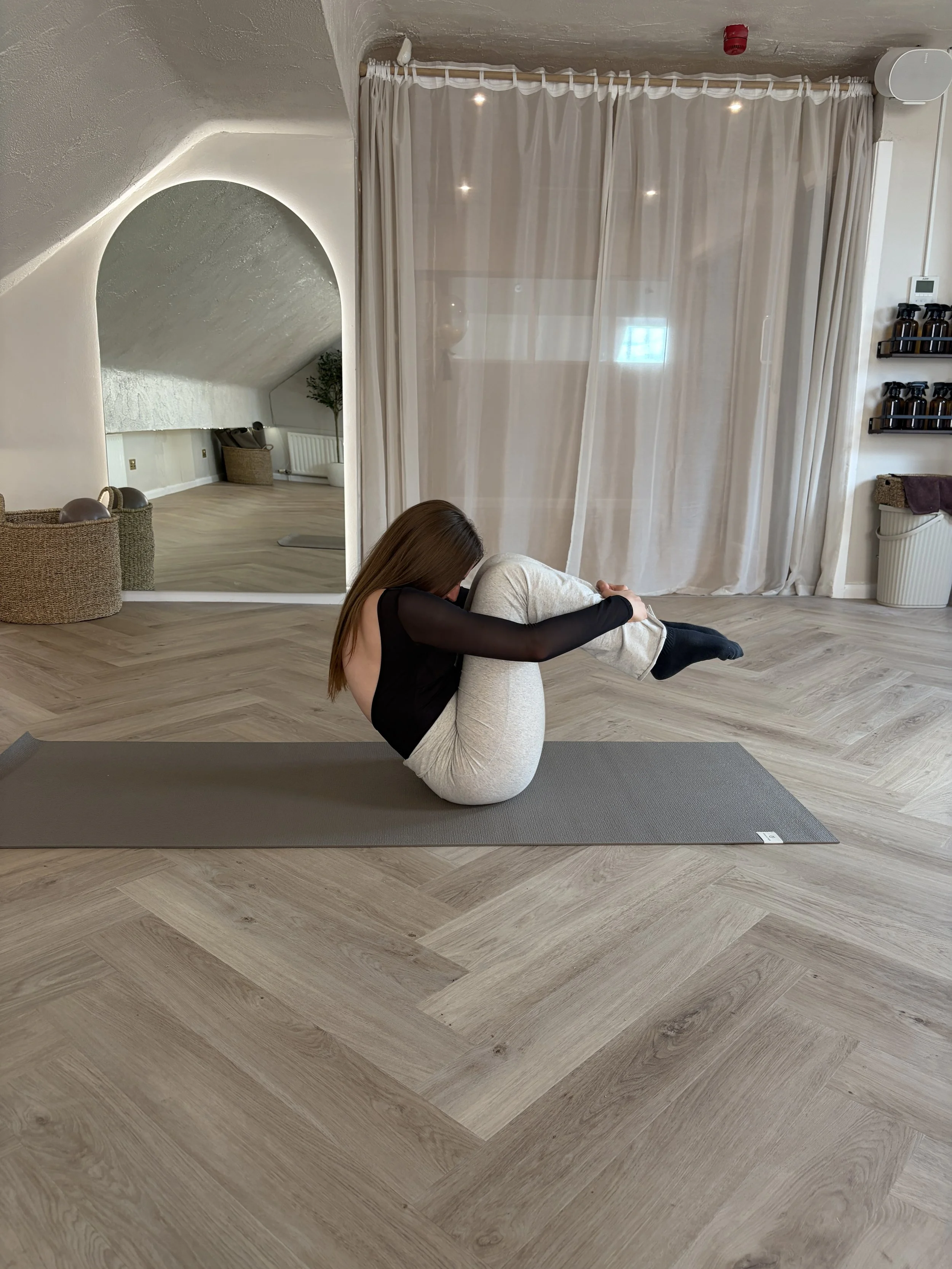 Woman in activewear performing a stretching exercise on a Pilates reformer machine in a modern, well-lit fitness studio with plants and mirrors.
