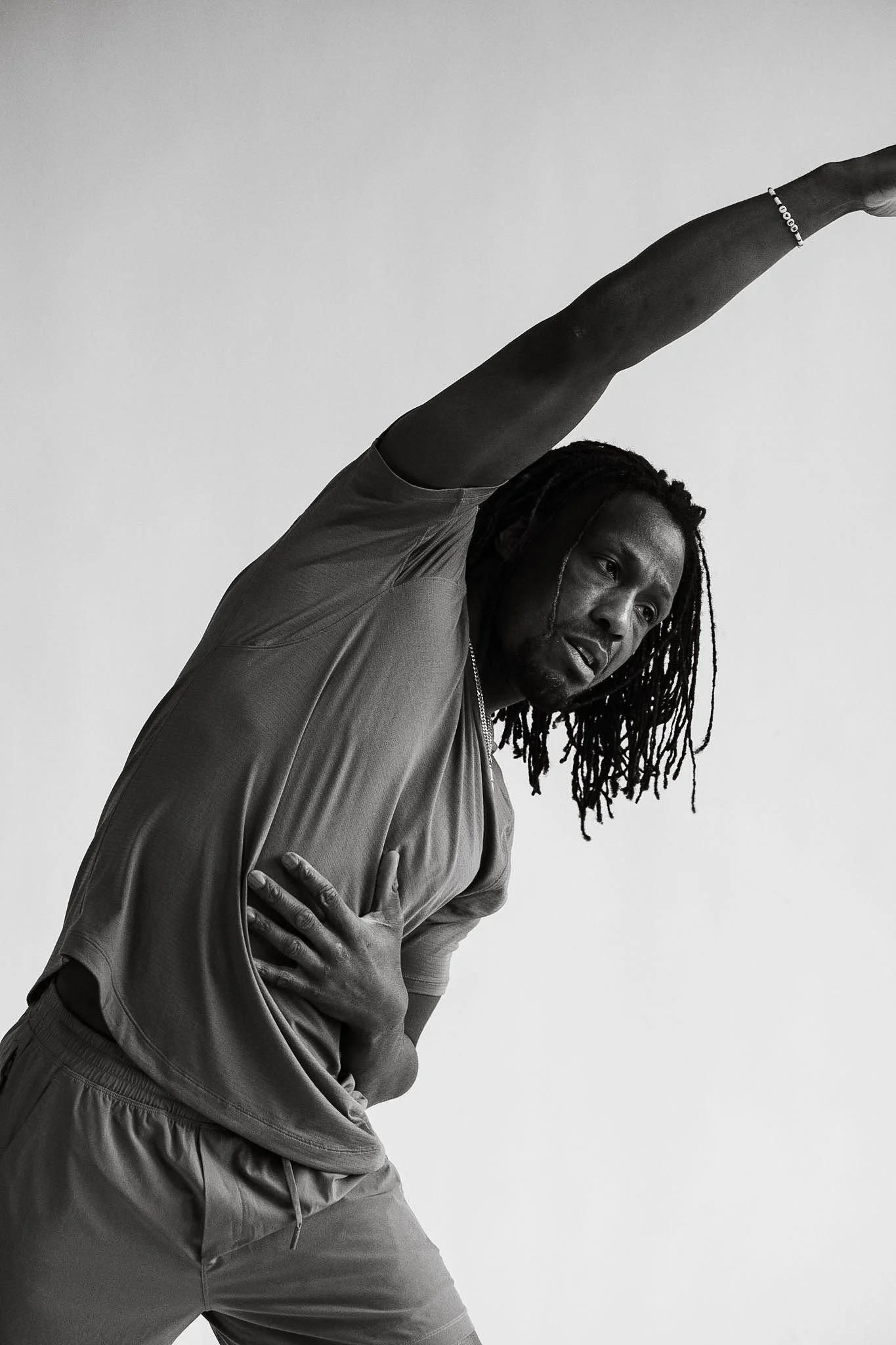A man with dreadlocks leaning to the side with one arm extended overhead and the other hand on his chest, in a stretching pose.