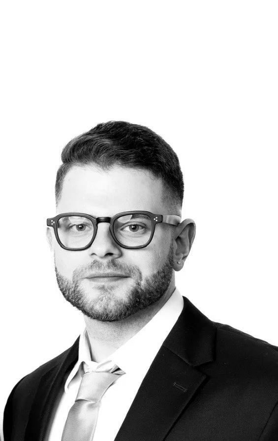 Black and white portrait of a man wearing glasses, a suit, and a tie, looking at the camera with a neutral expression.