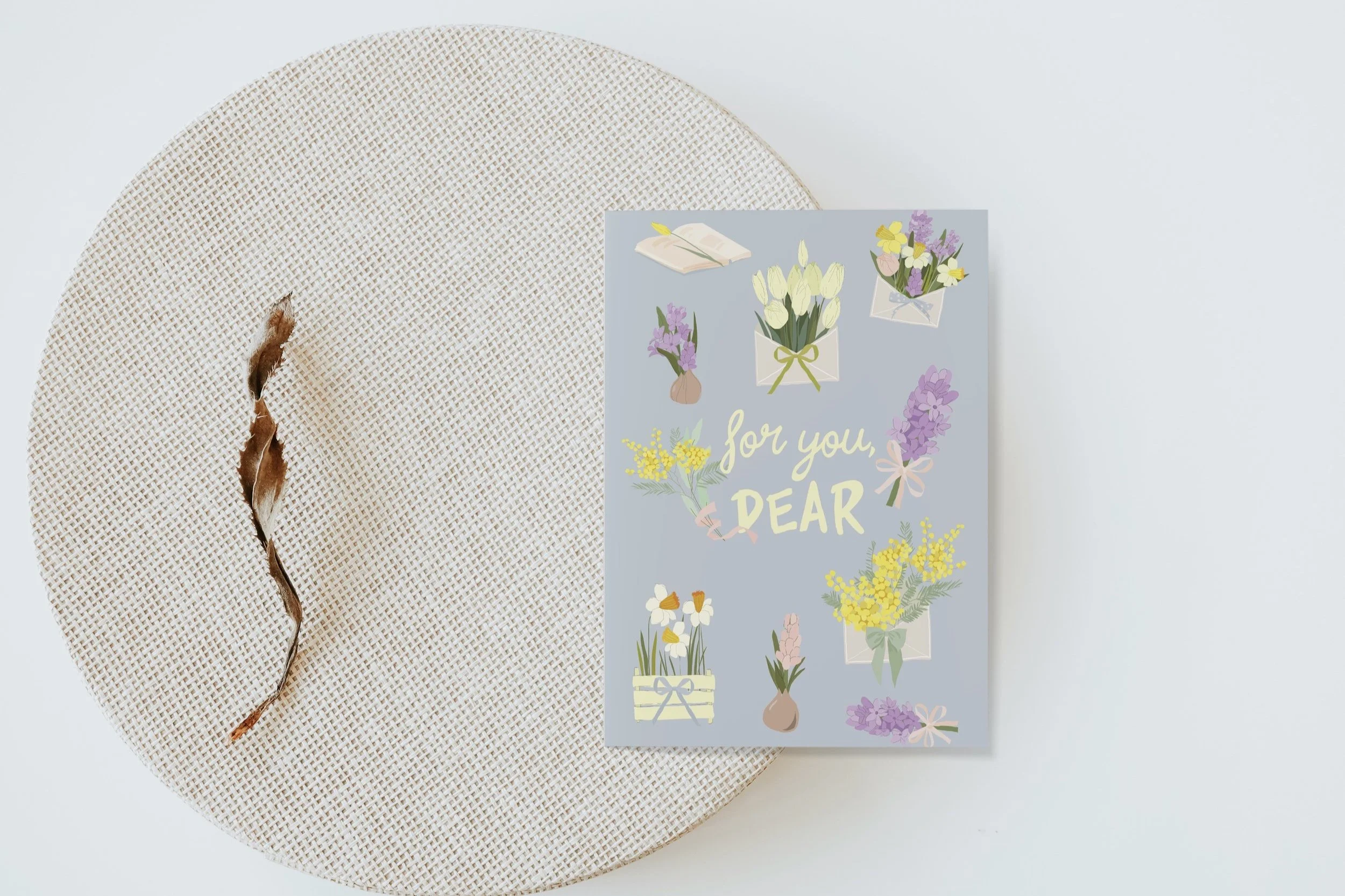 A greeting card with floral illustrations and the message "for you DEAR" on a white surface, next to a beige woven placemat with a torn decorative thread.