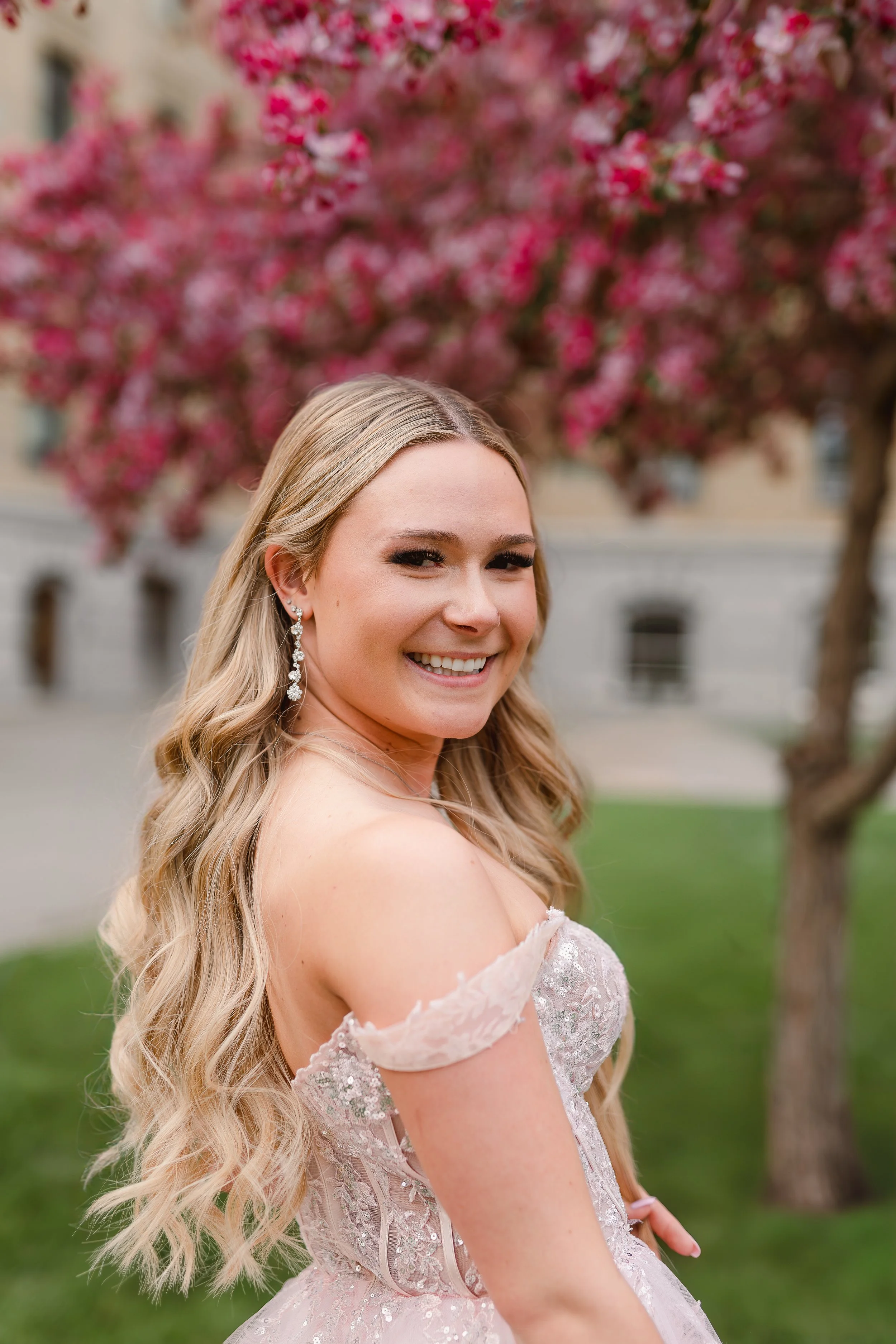 A smiling woman with long, wavy blonde hair wearing a pink, off-the-shoulder lace dress and dangling earrings, standing outdoors near a pink flowering tree.