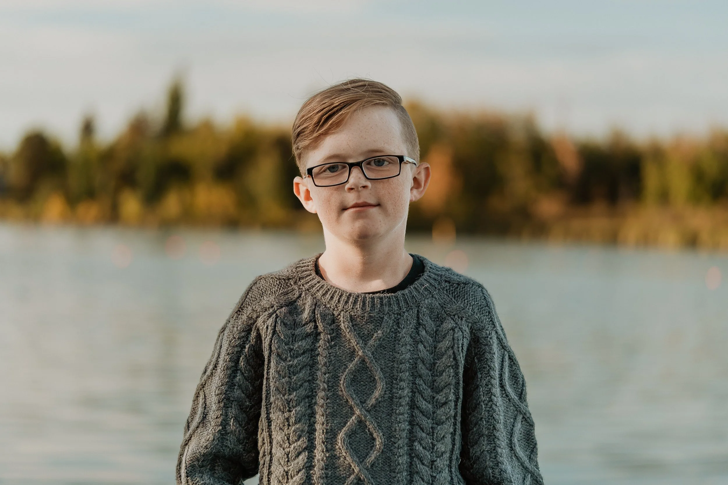 A young boy with glasses and a cable-knit sweater standing outdoors near a body of water, with trees and a cloudy sky in the background.