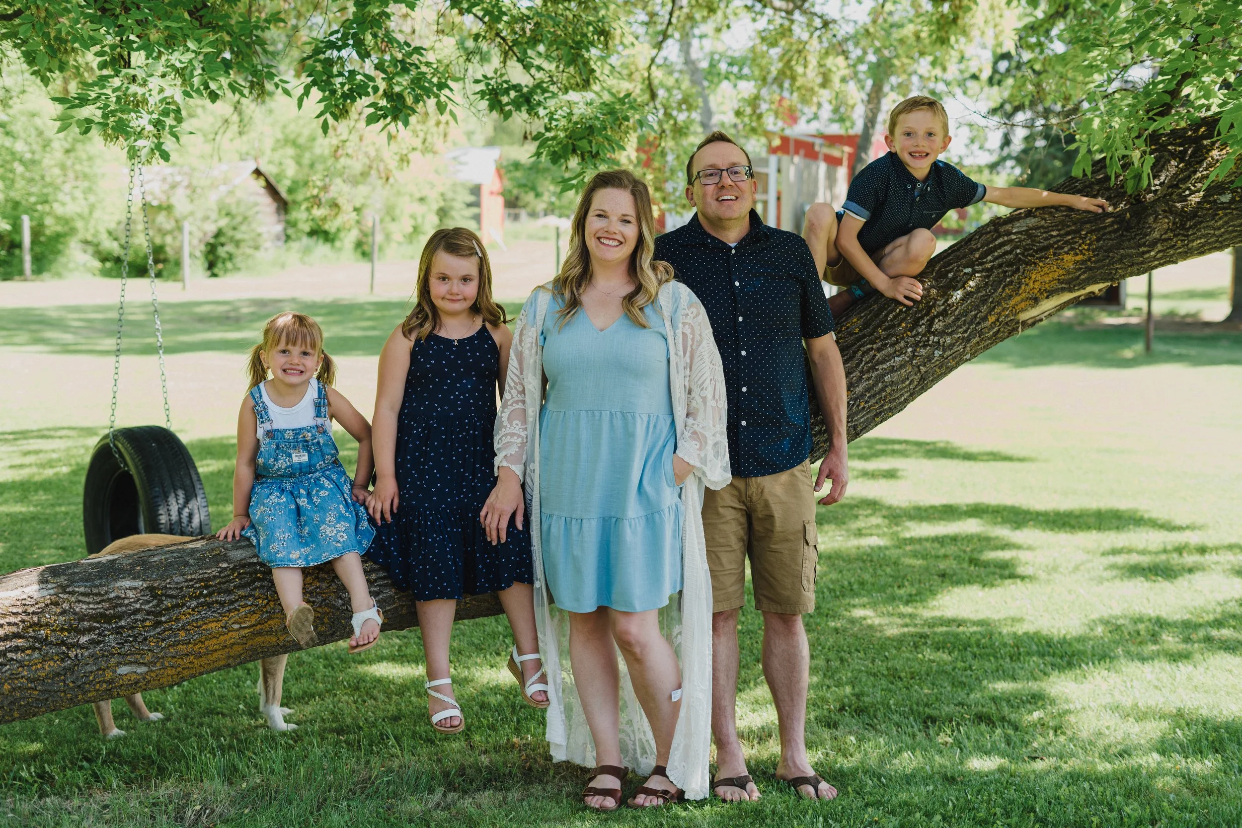 A family of six standing and sitting on a tree branch in a park with green grass and trees, smiling and enjoying a sunny day.