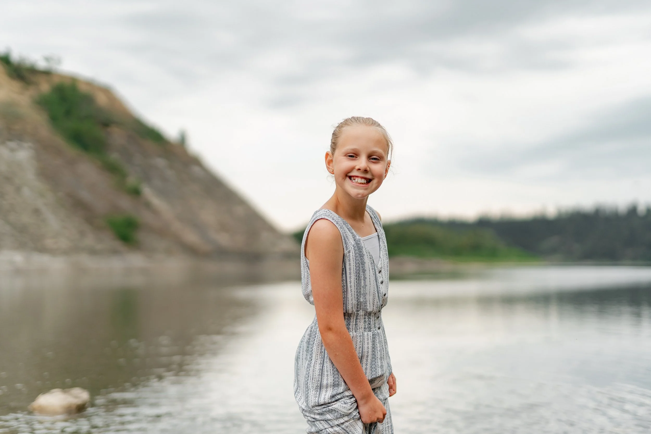 A young girl smiling and standing by a river with hills in the background.