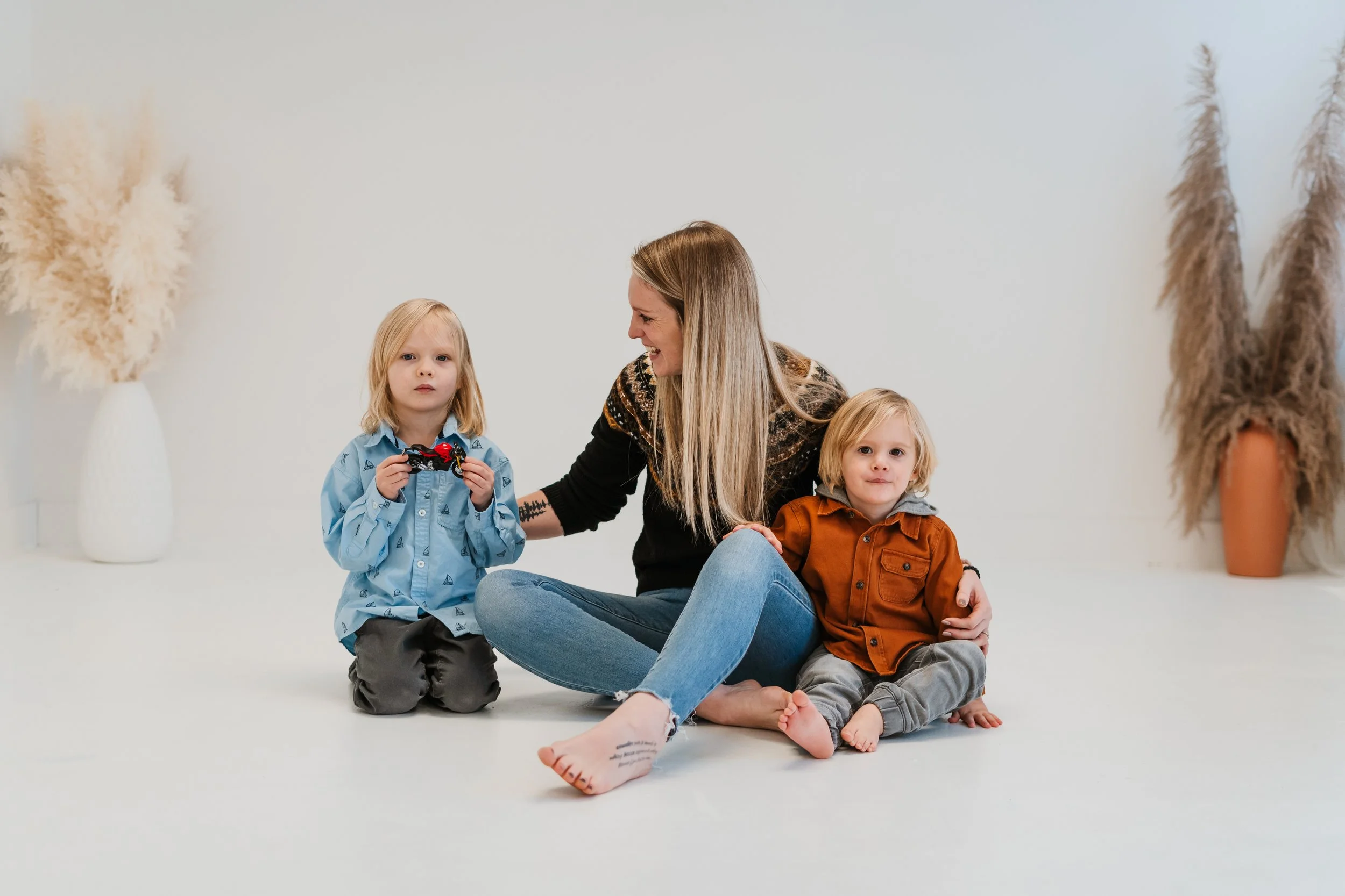 A woman sitting on the floor with two young children, one holding a toy car, in a minimalistic room with white walls and large vases with pampas grass