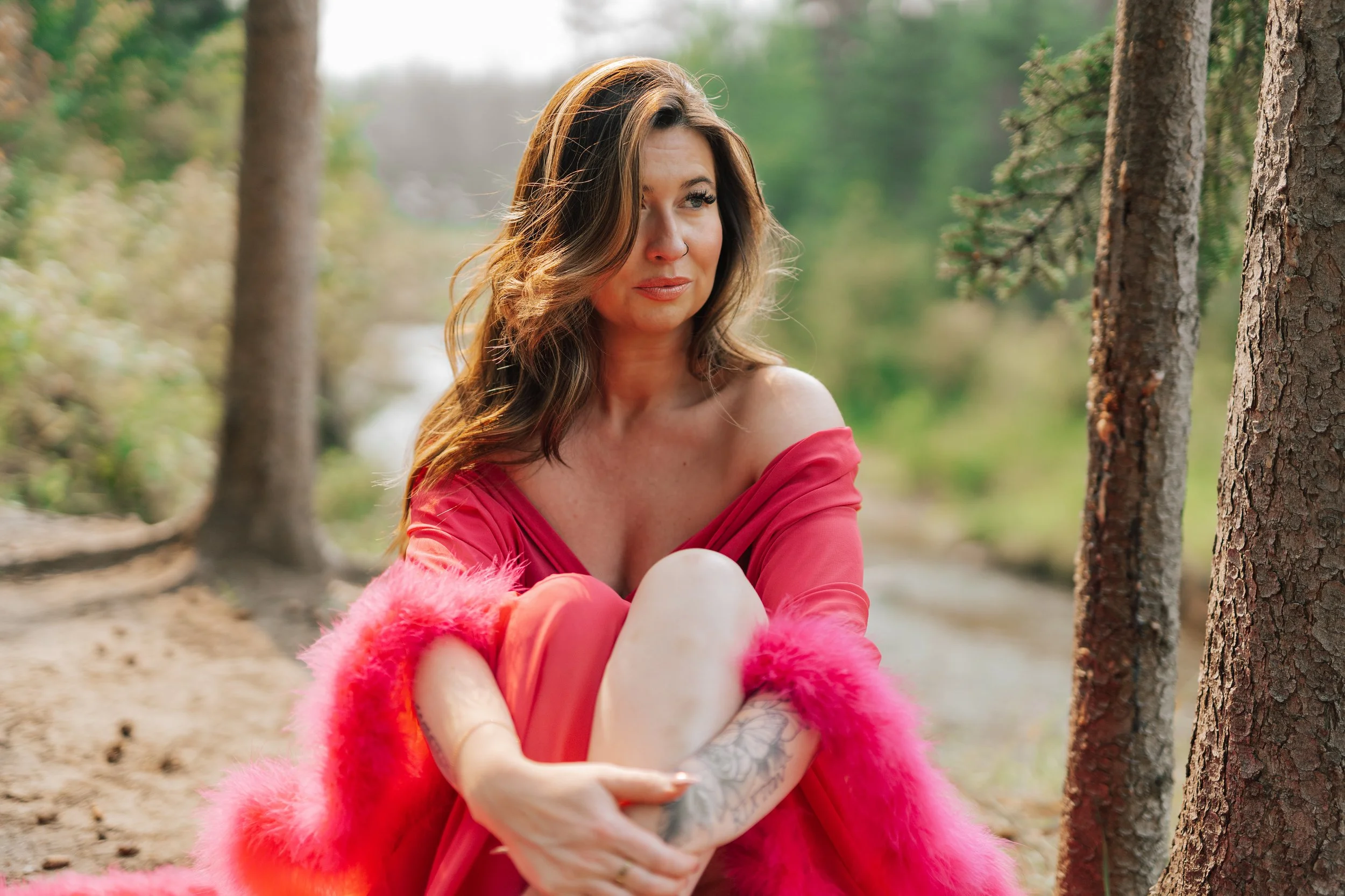 A woman with long, wavy brown hair sitting outdoors among trees, wearing a pink off-the-shoulder dress and a fluffy pink shawl.
