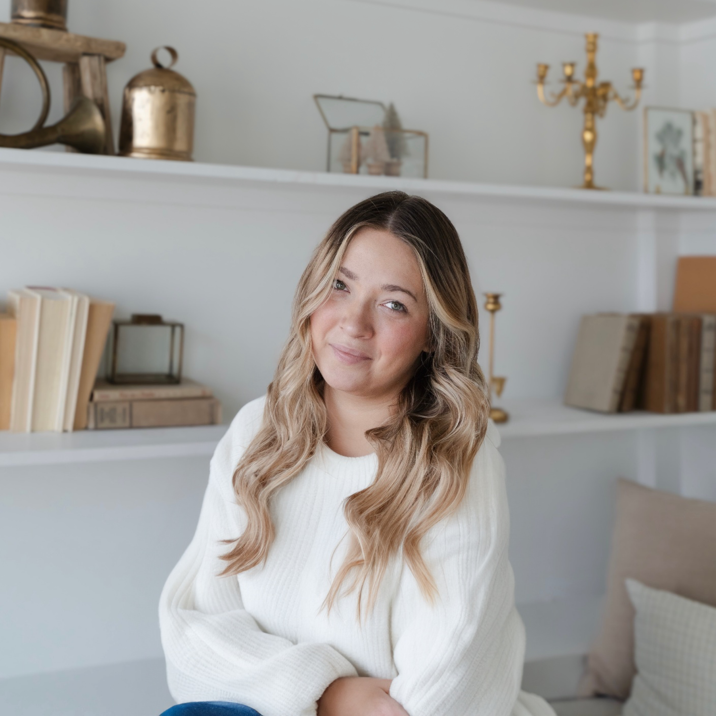 A woman with wavy blonde hair wearing a white sweater sitting on a beige couch in a bright room with white shelves filled with books, decorative objects, and gold accents.
