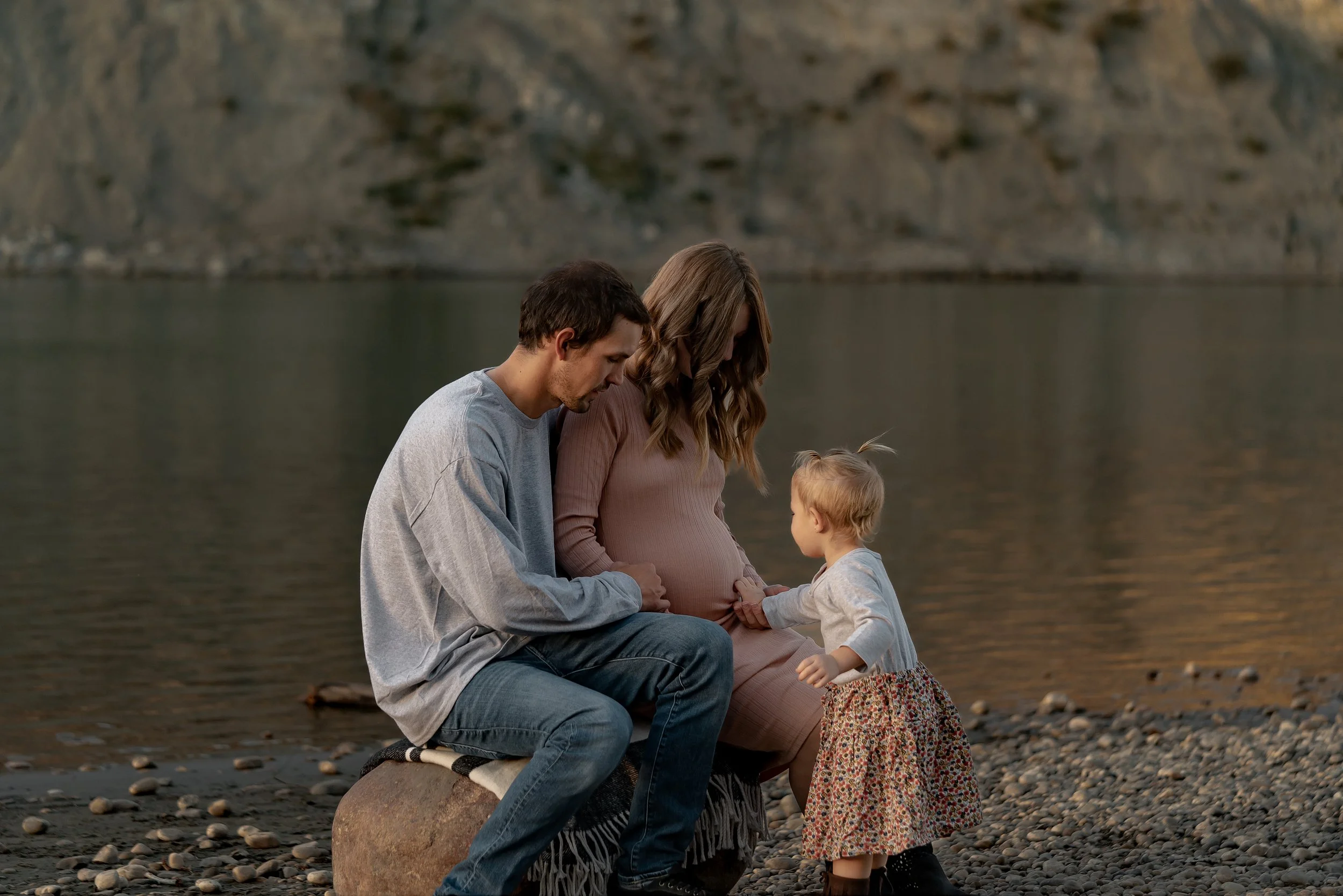 A family of three sitting by a lake with a pregnant woman, a man, and a young girl, during sunset.