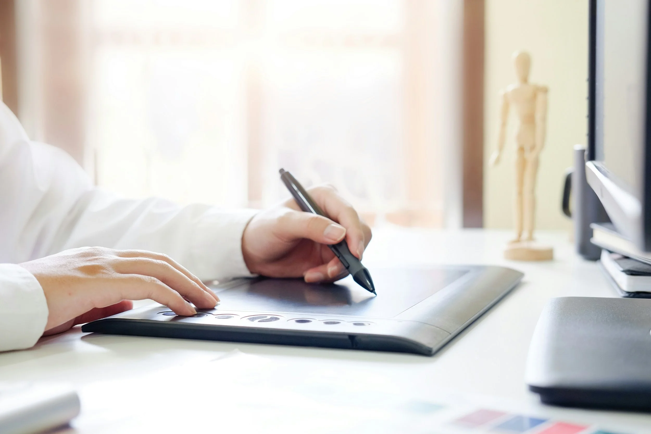 Person using a digital drawing tablet with a stylus on a white desk, with a wooden mannequin, a closed laptop, and a computer monitor in the background.