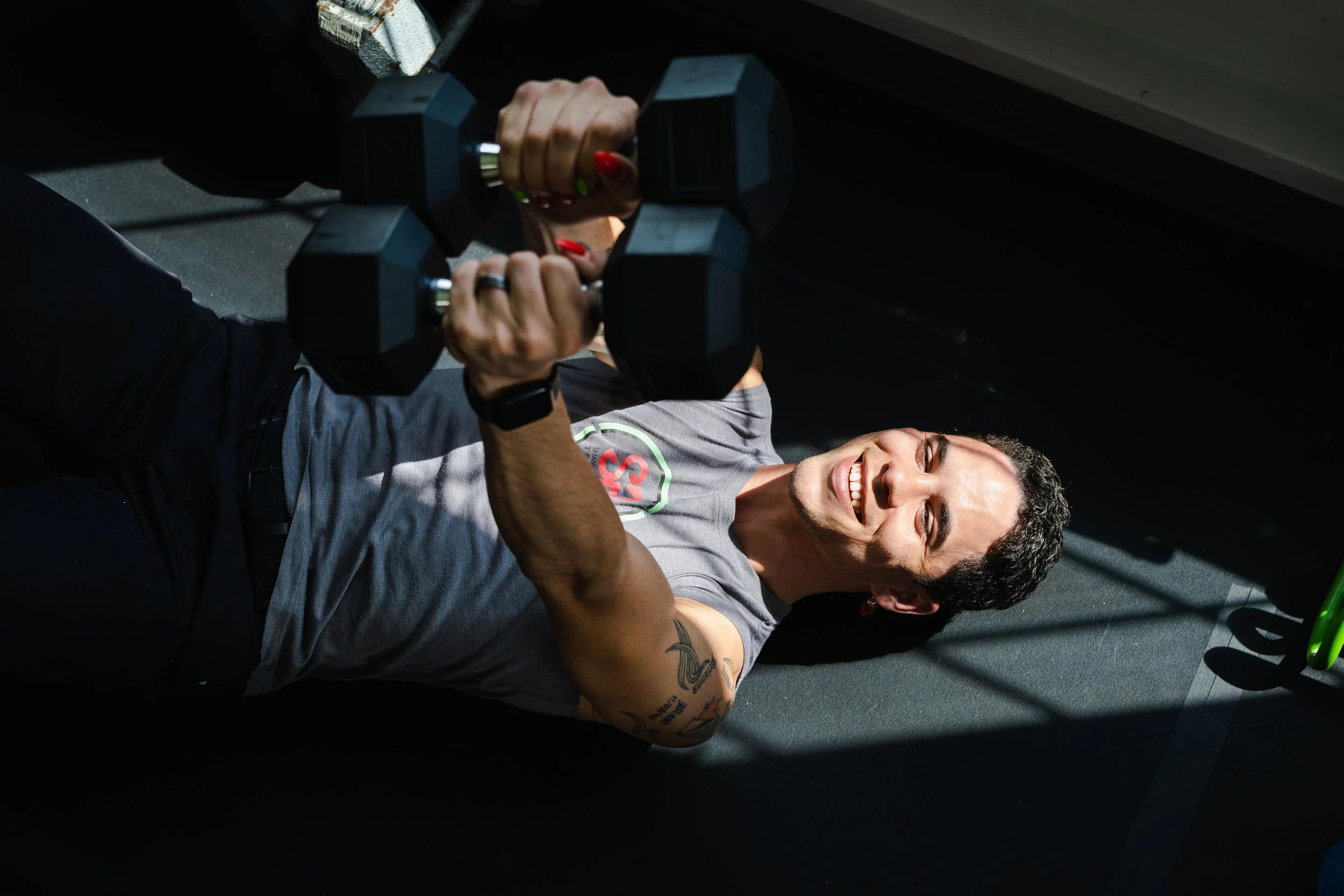A woman lying on the floor lifting dumbbells, smiling, wearing a gray T-shirt with a logo and black pants.