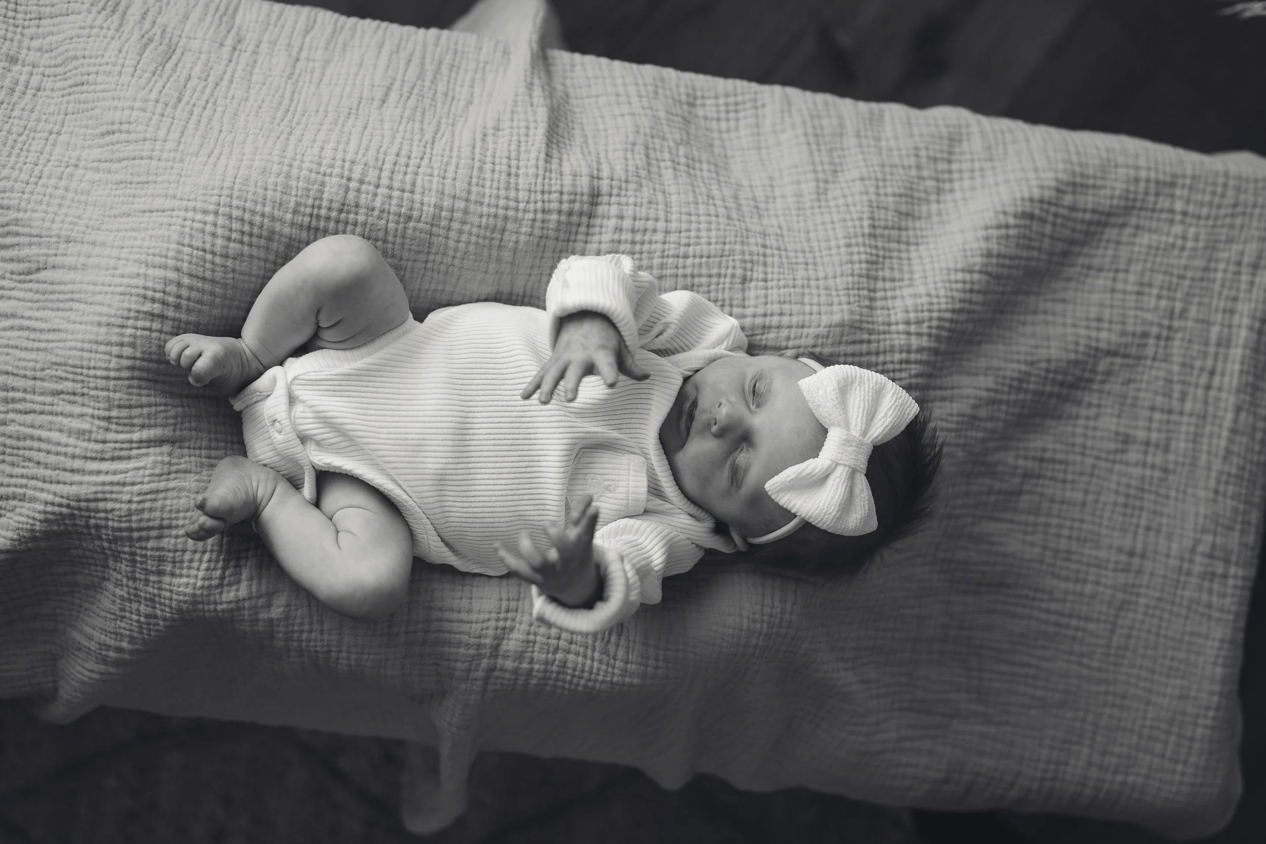 A baby girl with a bow headband sleeping on a textured blanket, dressed in a striped sleeveless onesie with long sleeves.
