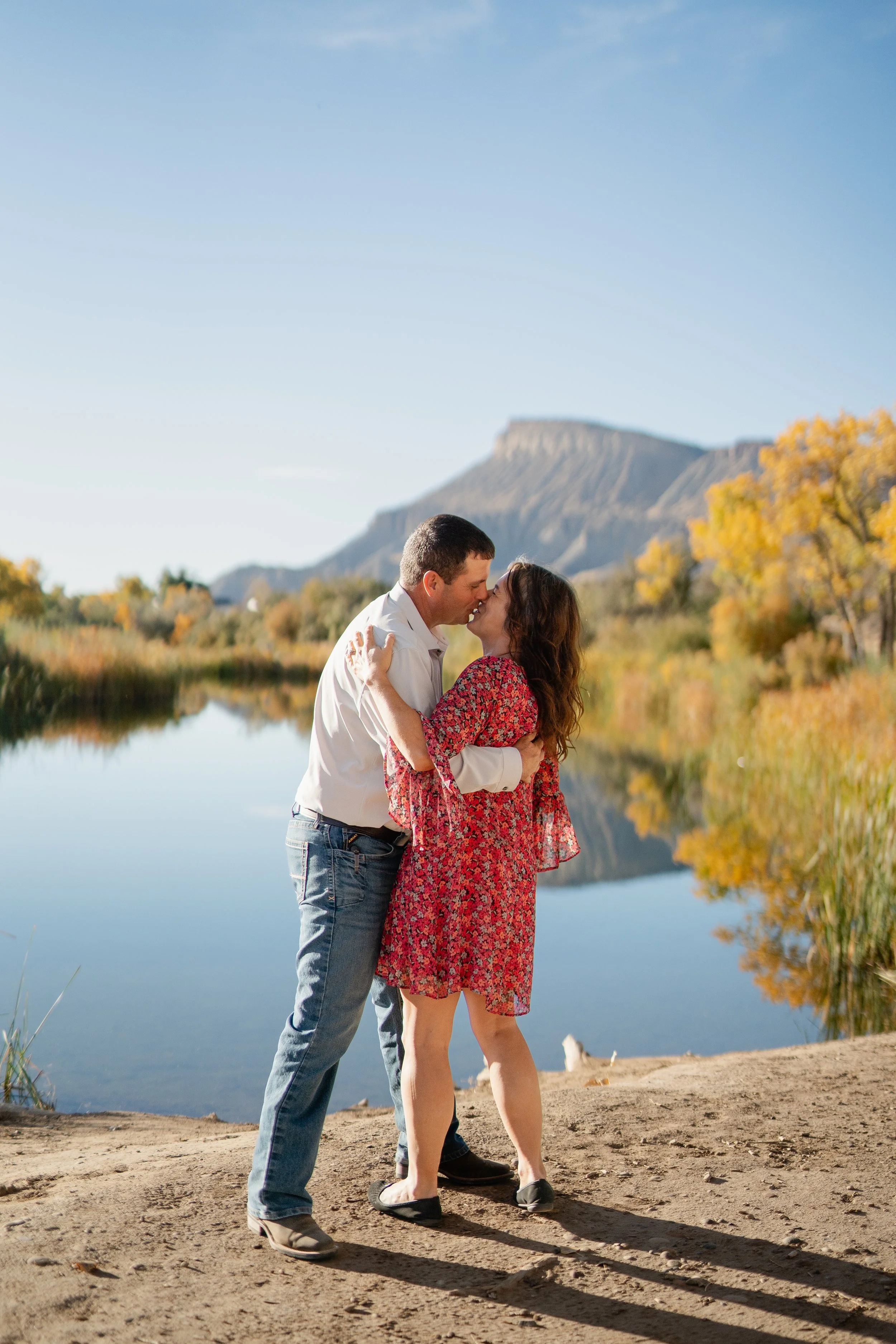 A couple kissing by a lake with autumn trees and mountains in the background.