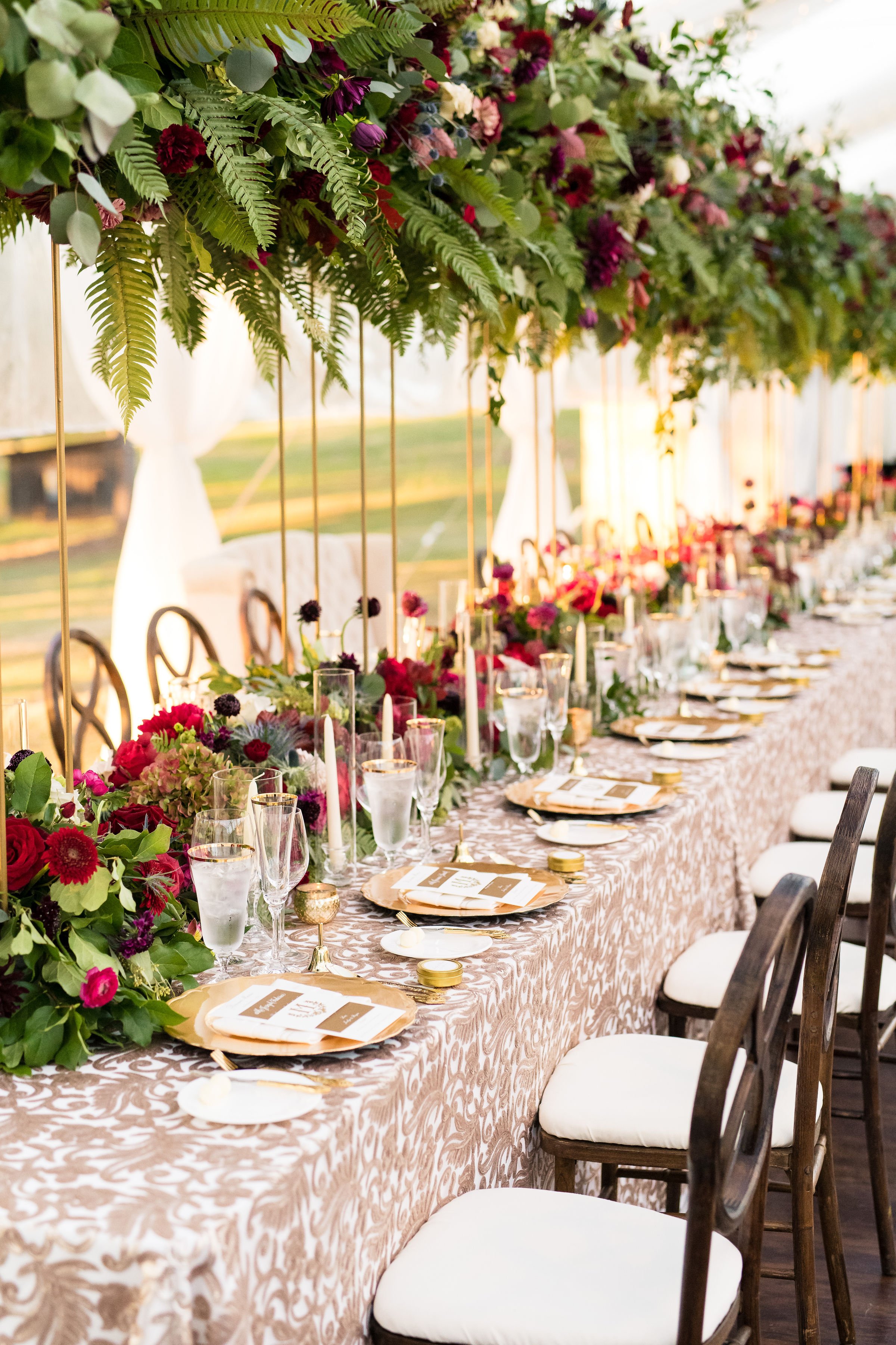 Elegant, long banquet table decorated with lush floral arrangements, candles, and table settings, set for a wedding or special event, with outdoor background and draped white curtains.