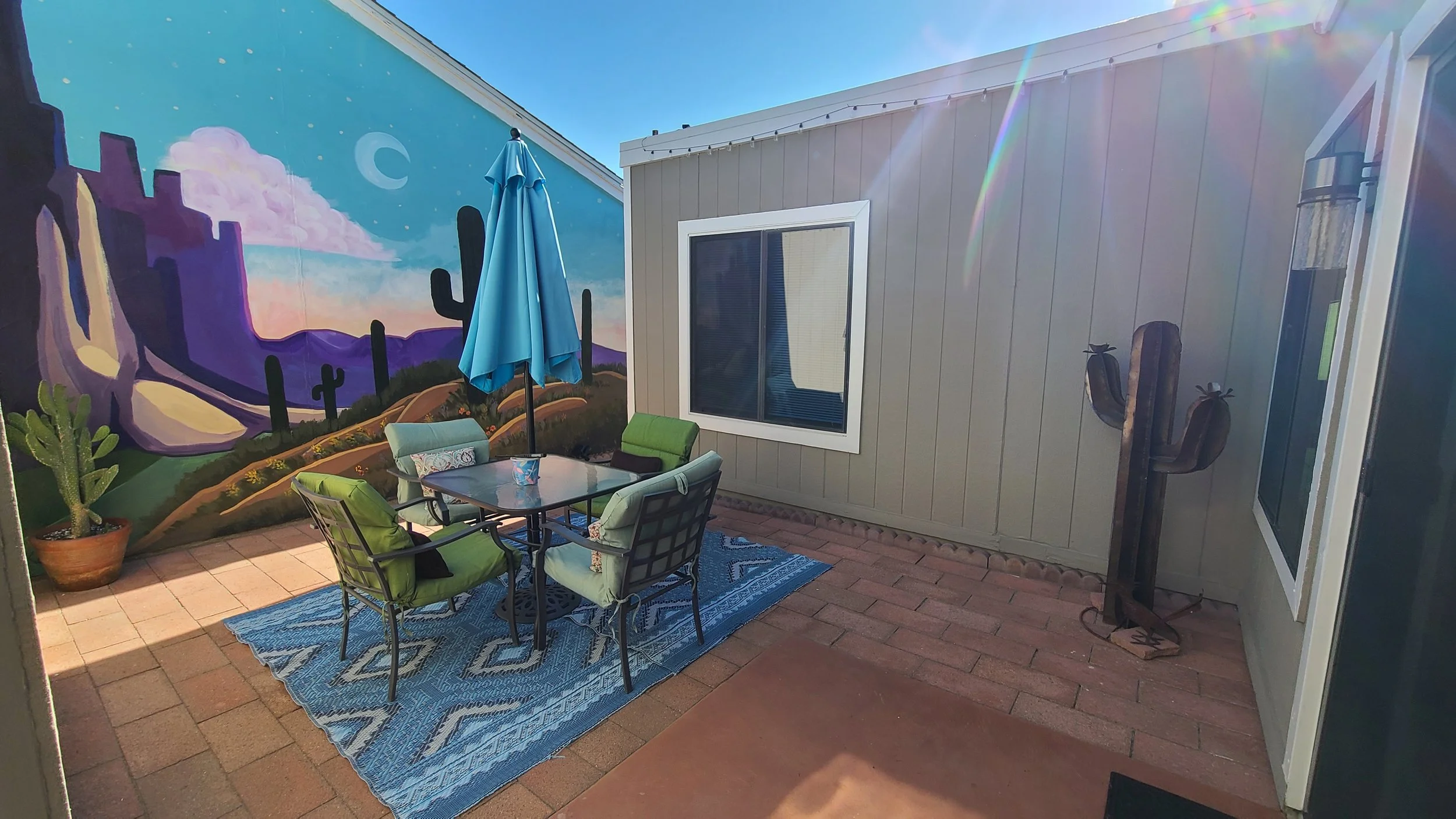 Outdoor patio with table, chairs, blue umbrella, cactus mural, and decorative cactus sculpture.