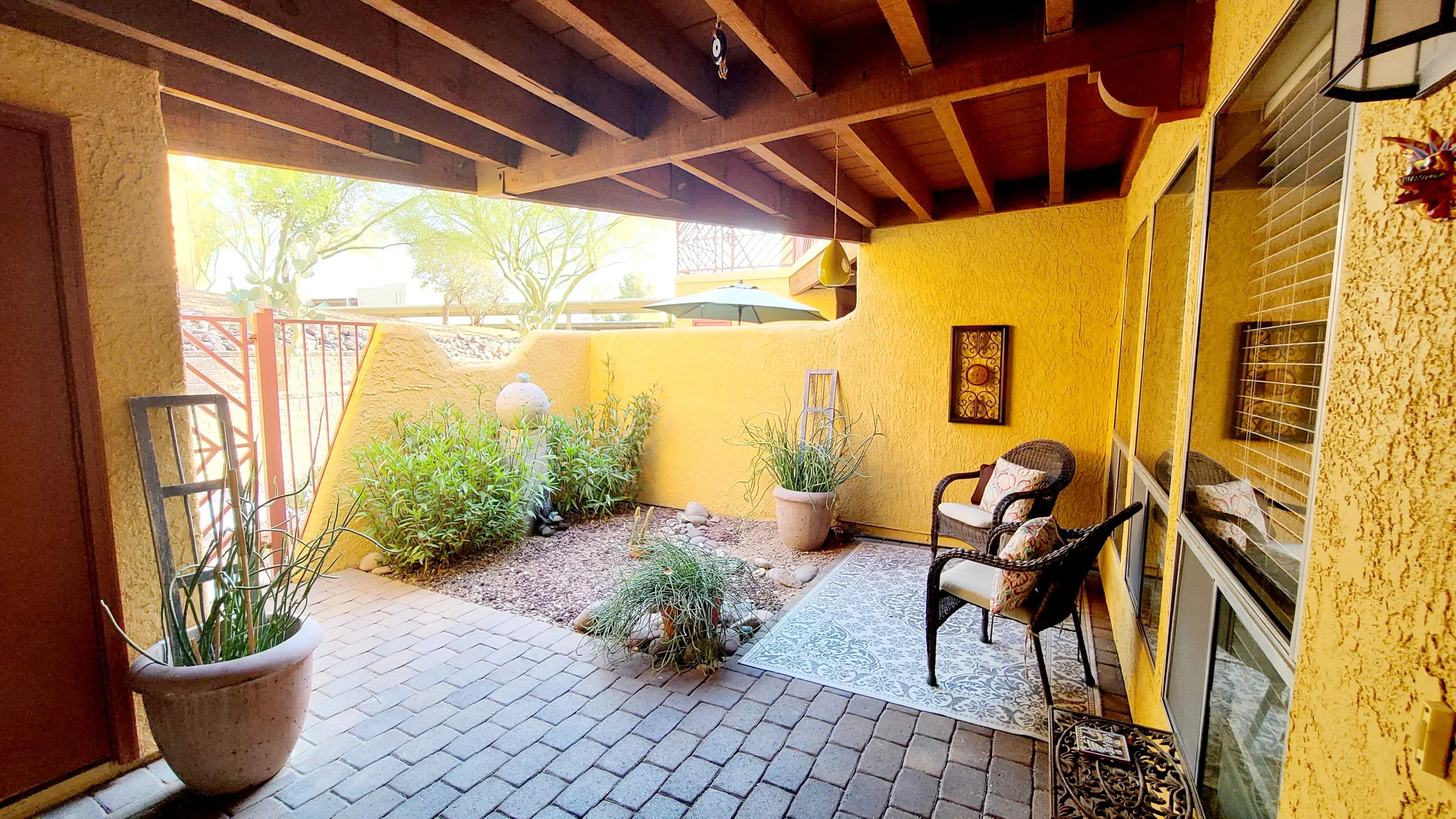 Small outdoor patio with yellow walls, wicker chairs, potted plants, and a decorative wall hanging under a wooden pergola.