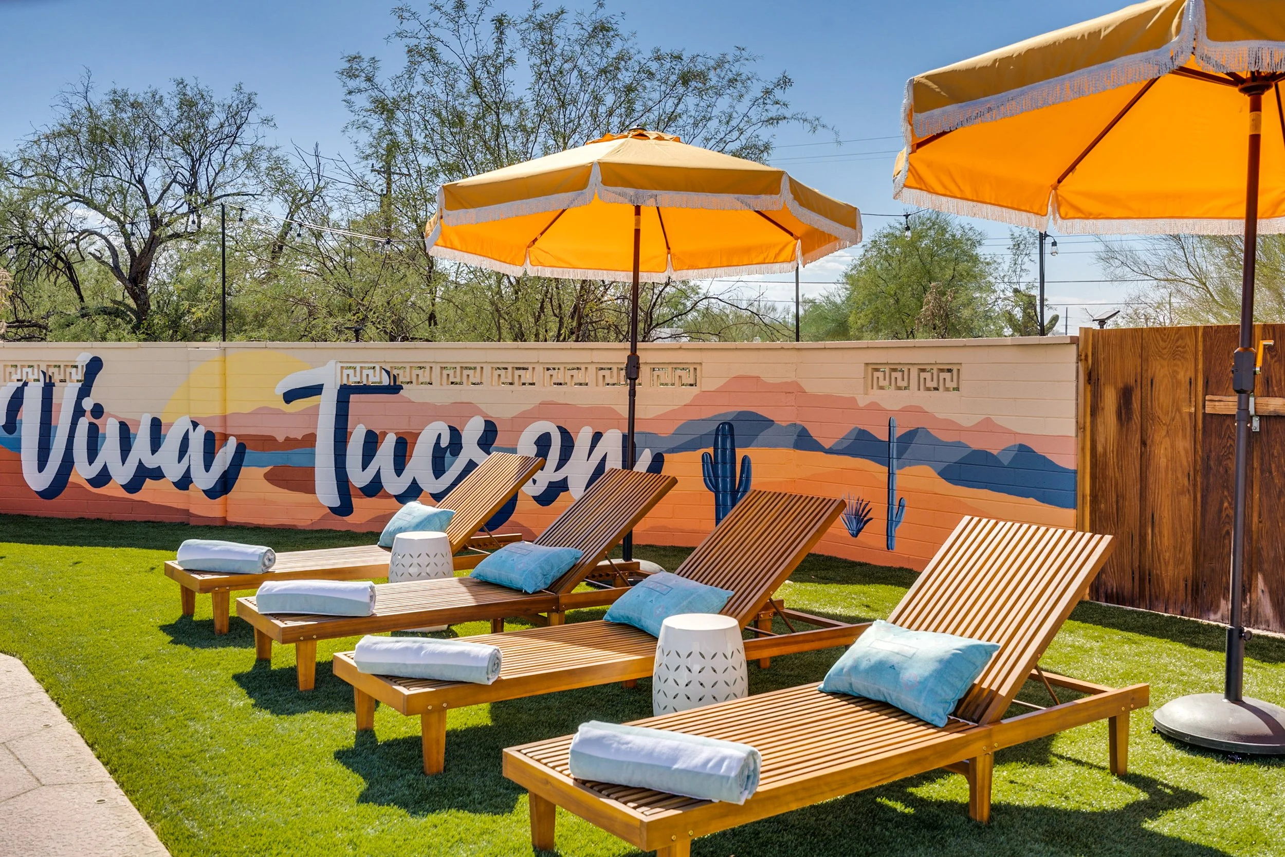 Outdoor lounge area with wooden loungers, yellow umbrellas, and a mural saying "Viva Tucson" with desert and mountain artwork.