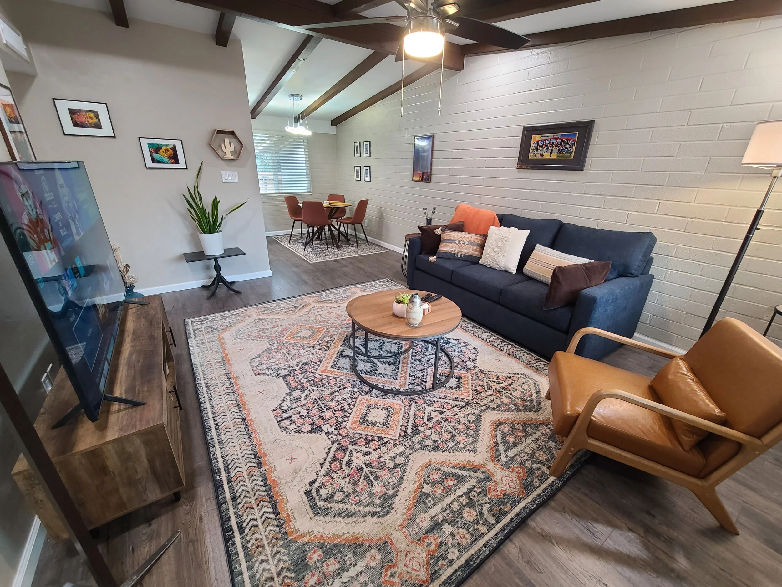 Cozy living room with a blue couch, brown armchair, and wooden coffee table on a patterned rug. Walls feature artwork and a plant. Visible TV and dining area.