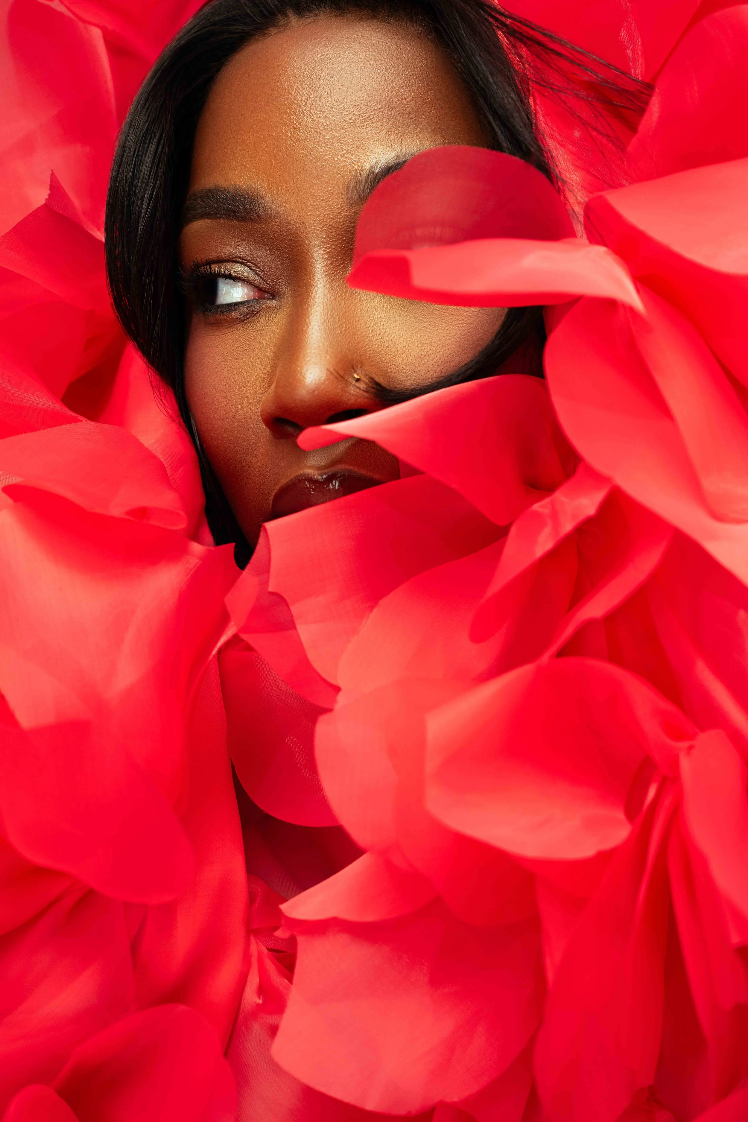 A close-up of a woman with dark hair and a small nose piercing, surrounded by large, bright red flower petals, with one petal covering her right eye.