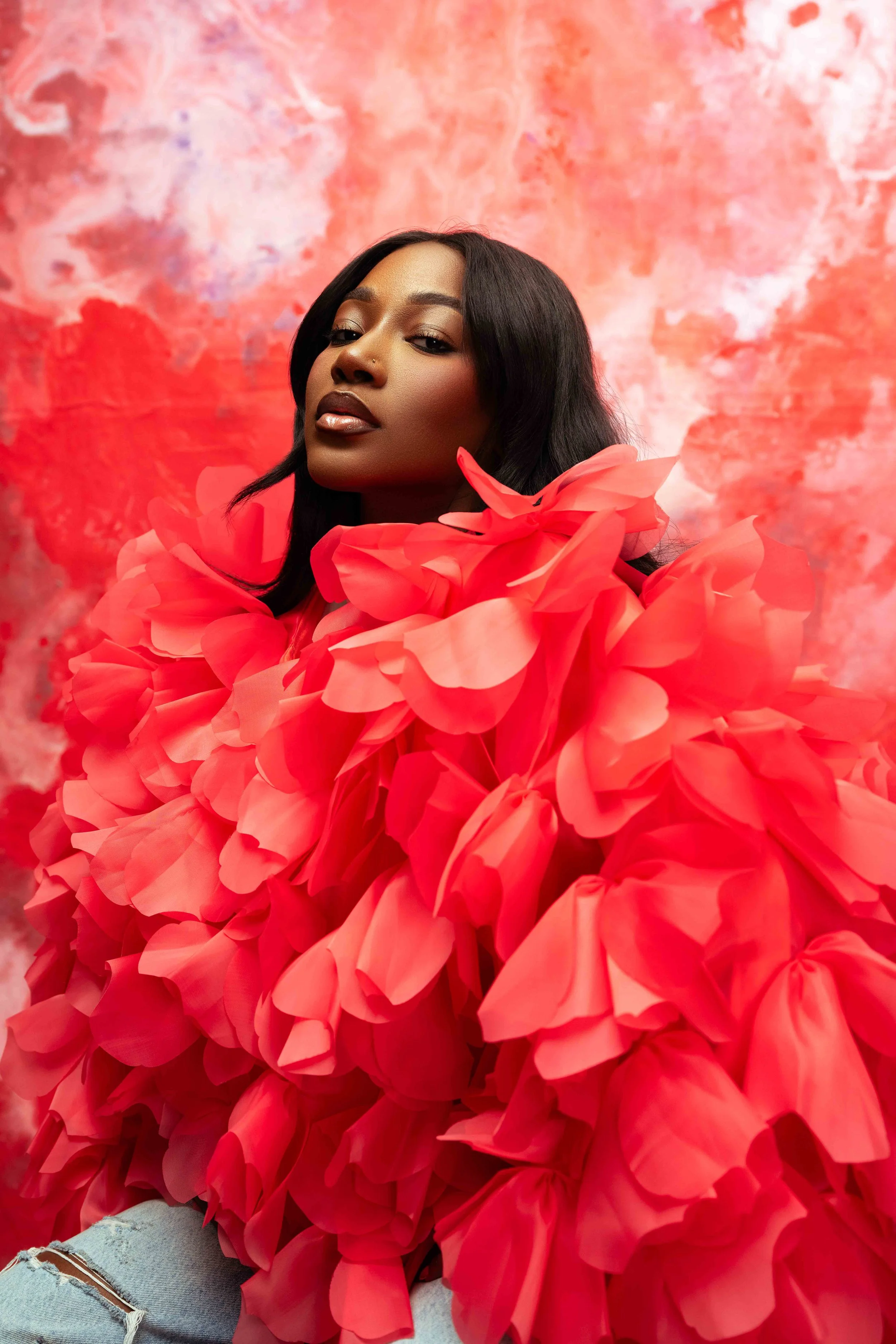 A woman with dark hair, wearing a bright red dress made of large, ruffled fabric flowers, posed against a pink and red abstract background.