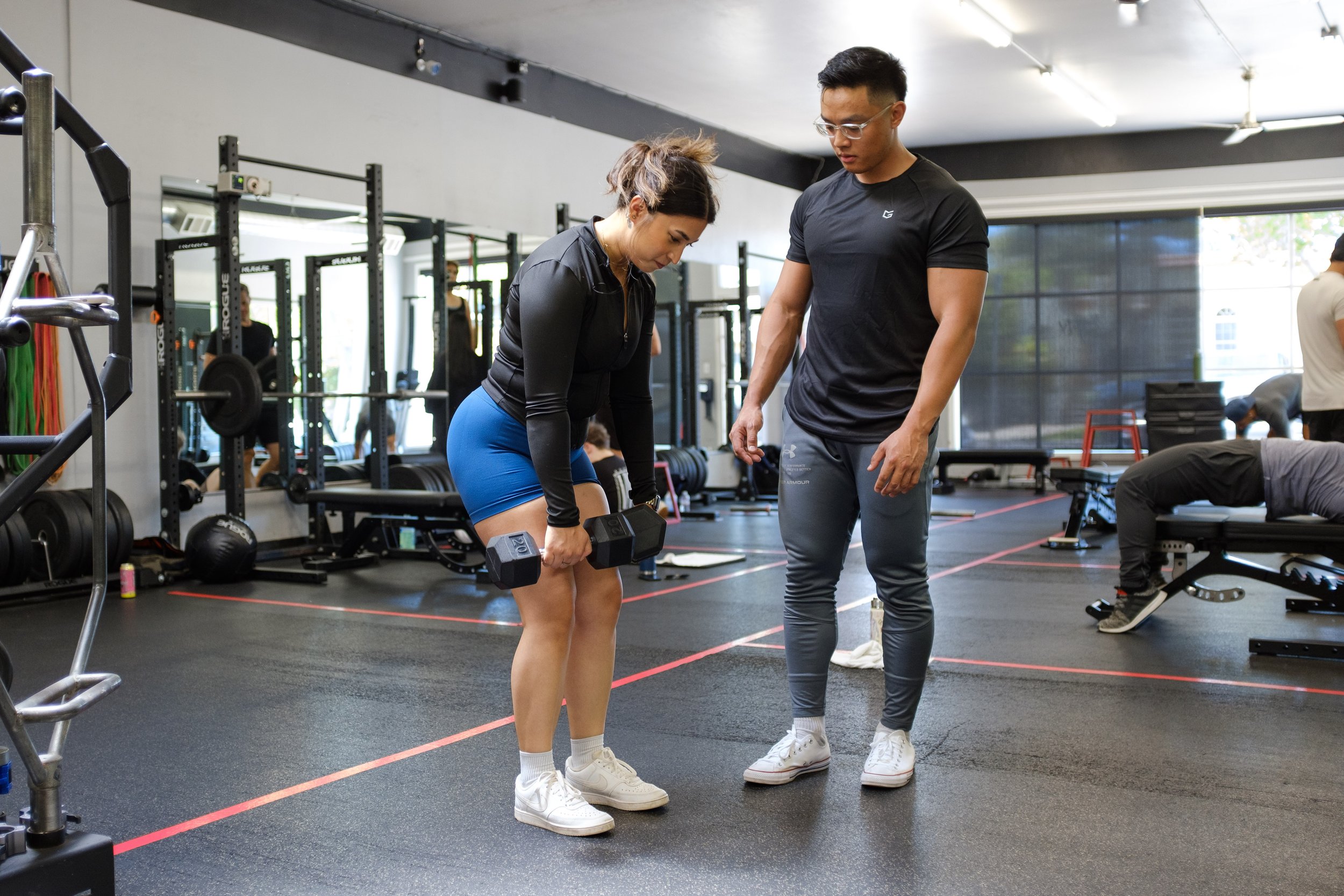 A woman practicing a dumbbell exercise with a trainer in a gym, with other people working out in the background.