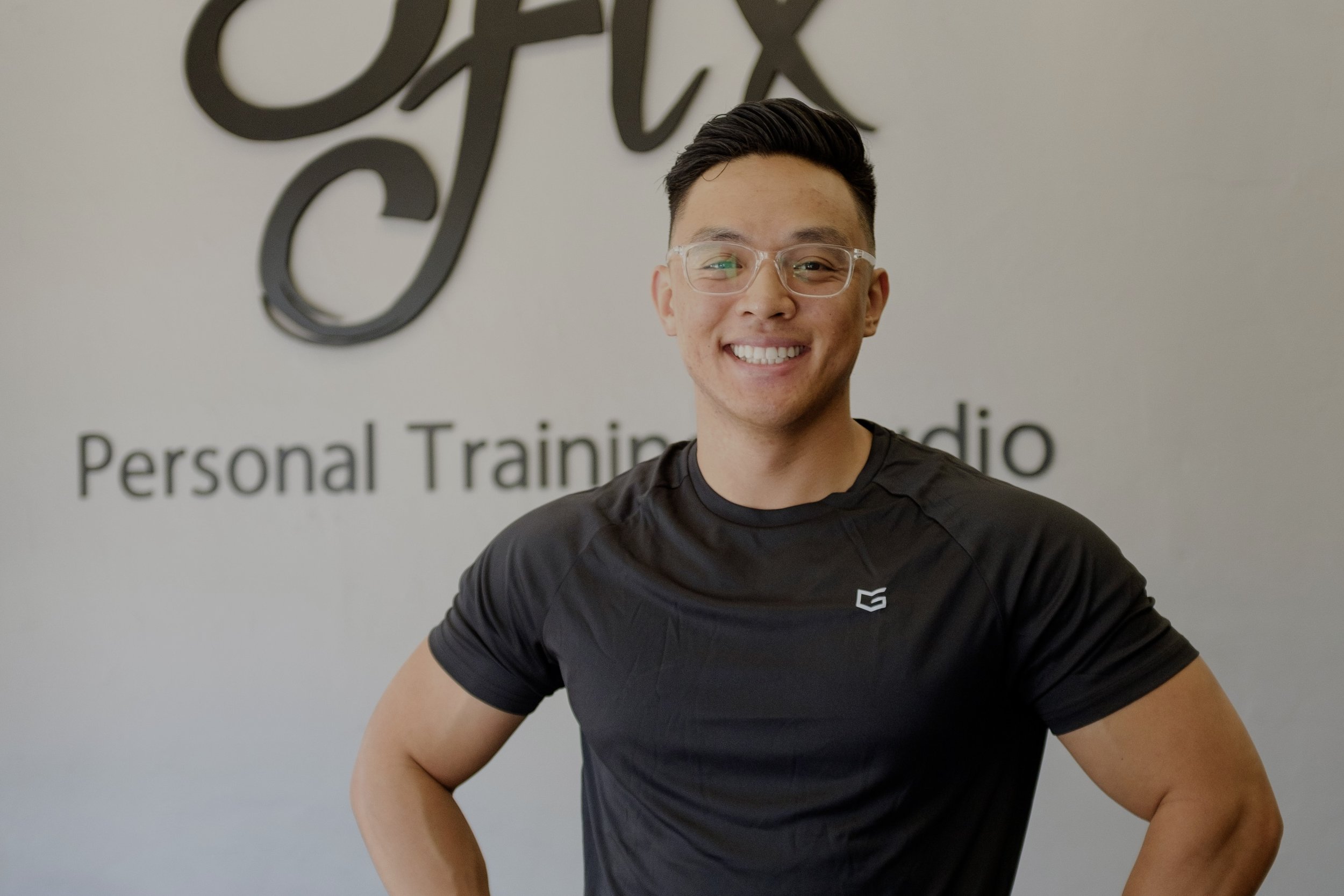 Smiling young man wearing glasses and a black athletic shirt standing in front of a wall with the words "Personal Training Studio" and a stylized logo.