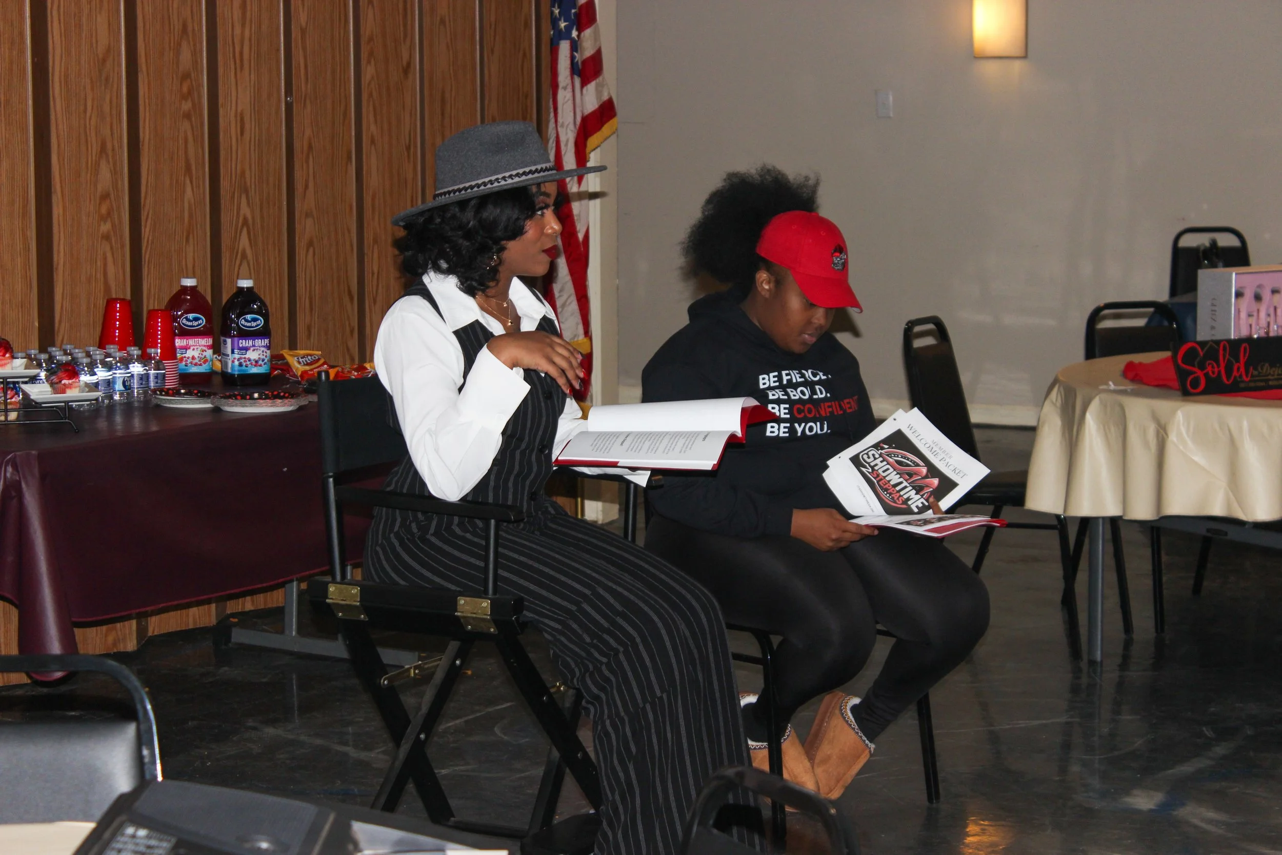 Two women sitting indoors, reading booklets. One woman is wearing a pinstripe suit and gray fedora hat; the other is dressed in a black hoodie and red cap. There are tables with snacks and drinks behind them, and an American flag in the background.