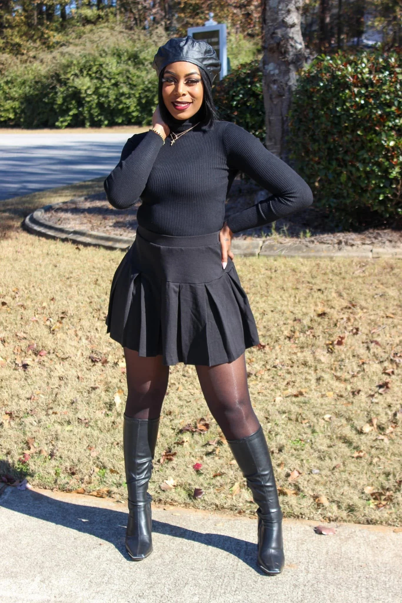 A woman standing outdoors on a sunny day, wearing a black sweater, black skirt, black stockings, and black tall boots. She has short black hair, and is wearing a black leather beret and jewelry. She strikes a confident pose with one hand on her hip and the other touching her neck, in front of a green shrub and tree.
