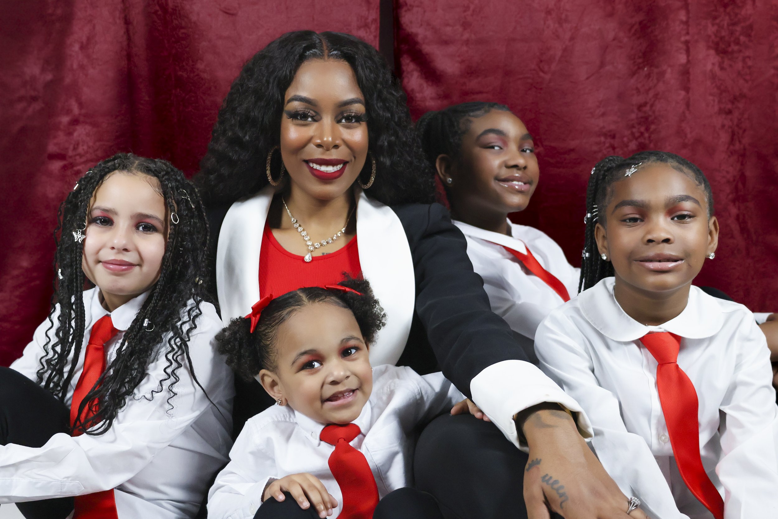 A woman with dark, curly hair and a bright red lipstick, dressed in a black blazer and white shirt, sitting with four young girls in white shirts with red ties, in front of a red curtain. The women and girls are smiling, with the women having jewelry