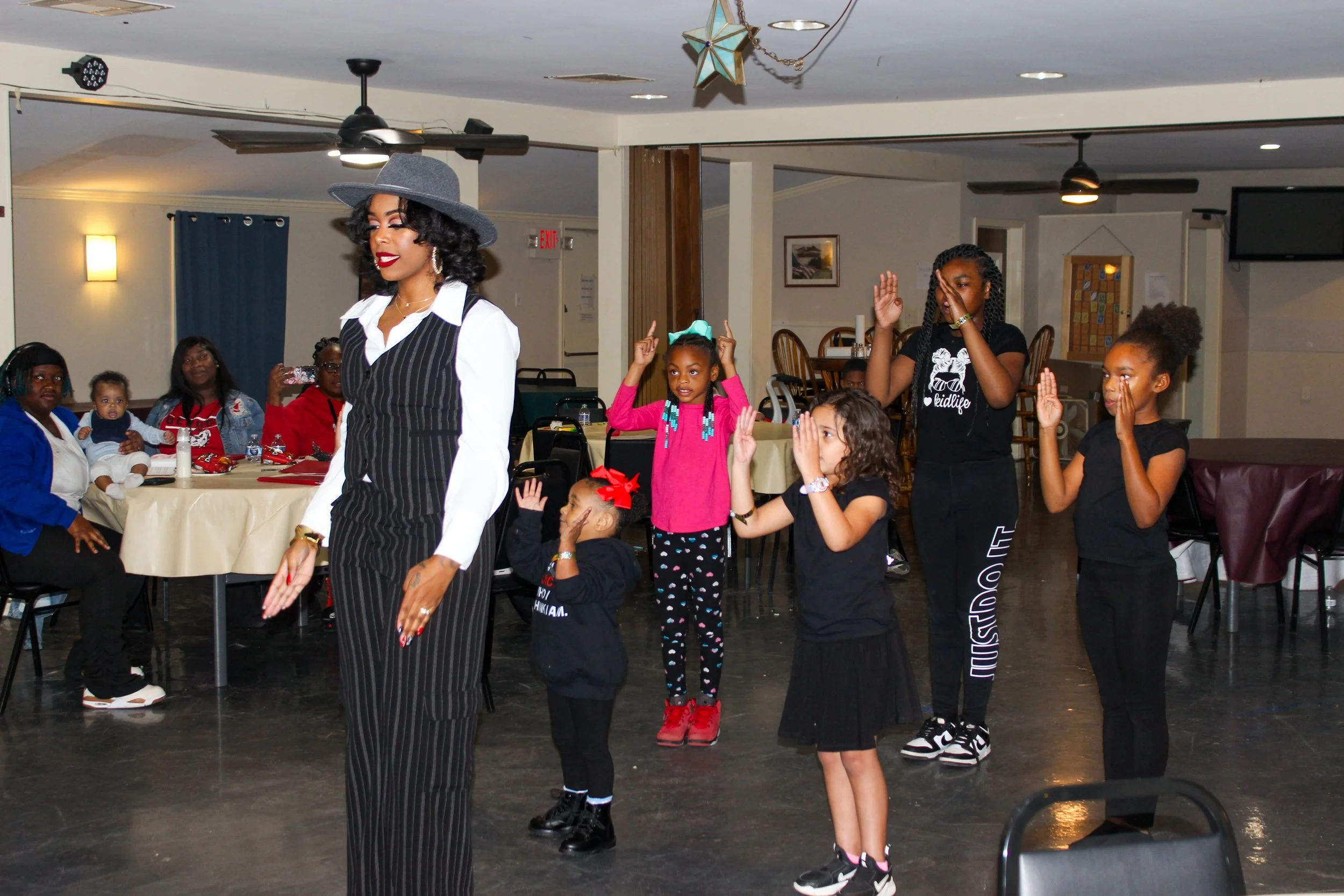A woman leading a dance or activity with children in a social gathering or party setting, some children have hands raised or covering their faces, with others sitting at tables in the background.